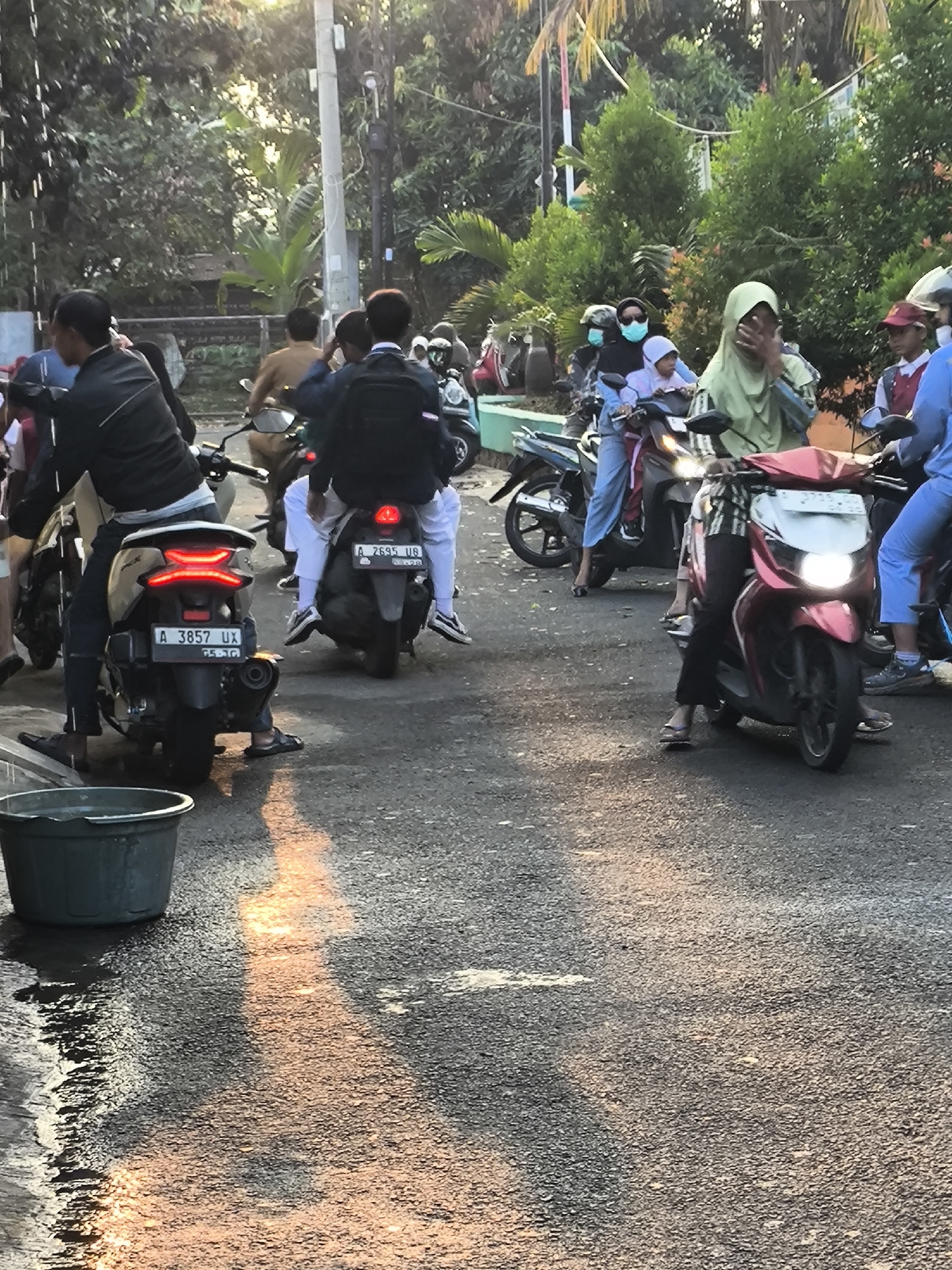 This is a medium-shot, outdoor, eye-level, full-color photograph of a street scene in Cilegon, Indonesia, likely during late afternoon due to the warm, directional lighting casting long shadows. The image is filled with several people on motorcycles, suggesting traffic or a gathering.
In the foreground, a dark gray plastic tub is visible on the left, partially filled with water, with reflections of light on the wet asphalt. The road surface itself is asphalt, showing wear and tear and patches of wetness.
The primary subjects are individuals on motorcycles. On the left, a person in a black jacket and black pants is on a motorcycle, facing away from the camera. To the right of this, another motorcycle is seen from behind, with its red taillight illuminated. The license plate on this motorcycle reads "A 3857 UX".
Further back and to the right, several other motorcycles and individuals are present. One motorcycle has a rider wearing a light green hijab and another person behind them wearing a lighter green hijab. Another motorcycle is partially visible, with a rider wearing a blue top and pink pants. In the background, more people on motorcycles and greenery from trees and bushes can be seen. The overall impression is a busy street with multiple motorbikes and passengers, possibly navigating through traffic or waiting.