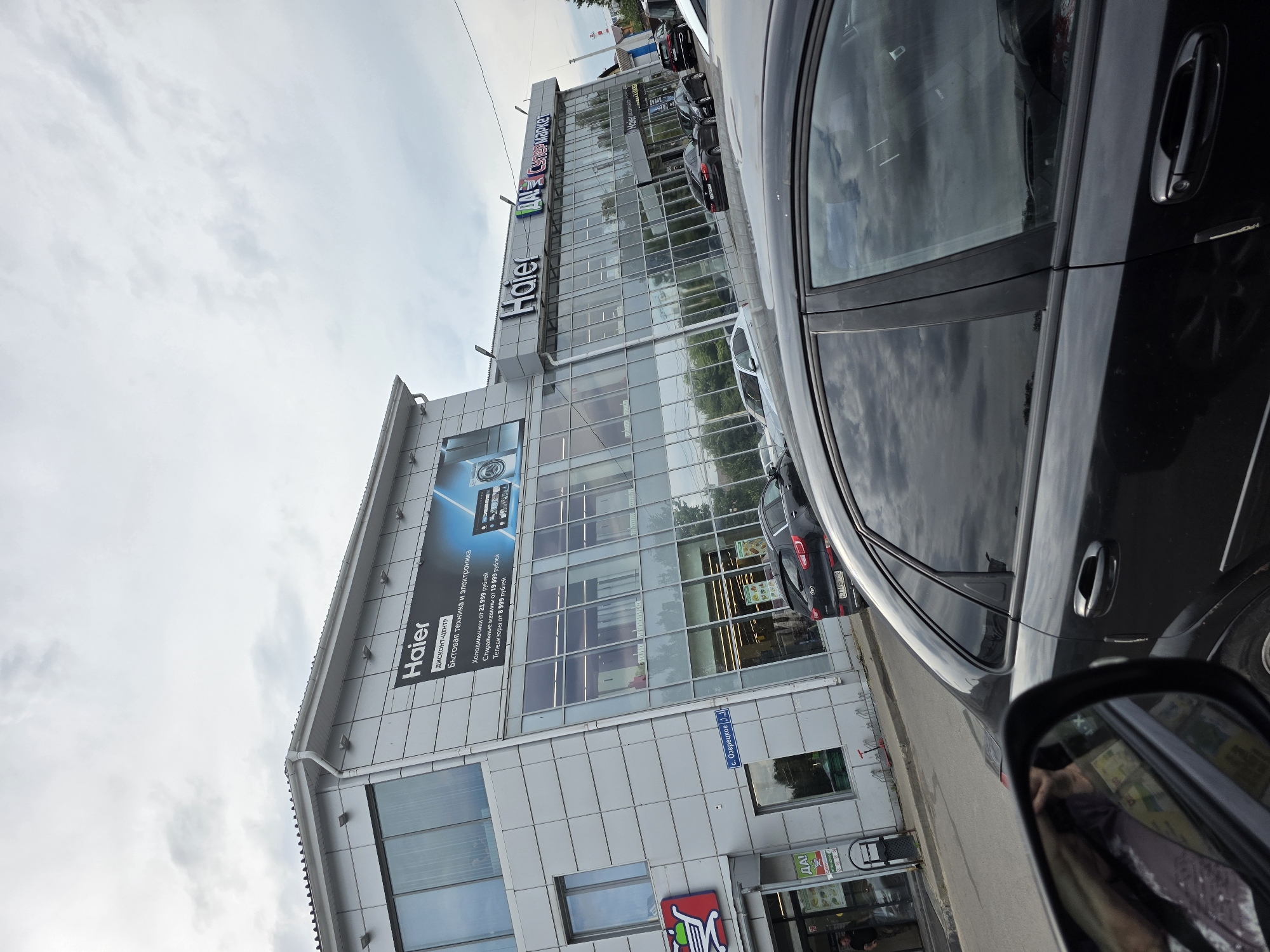 A wide-angle, eye-level outdoor shot captures the front of a Haier discount center and electronics store in the city of posyolok Sovkhoza "Ostan'kino", Russia. The building features a modern facade with large glass windows reflecting a cloudy sky. A large banner advertises Haier appliances, including washing machines and televisions. The store's name, "Haier", is prominently displayed above the entrance. To the left of the Haier store, a grocery store with a red shopping cart logo is visible. Several cars are parked in front of the building, and a black sedan is in the foreground, partially obscuring the view of the store. The weather appears to be overcast, with dramatic cloud formations in the sky. A street sign indicates "s. Ozeretskoe 3.1".