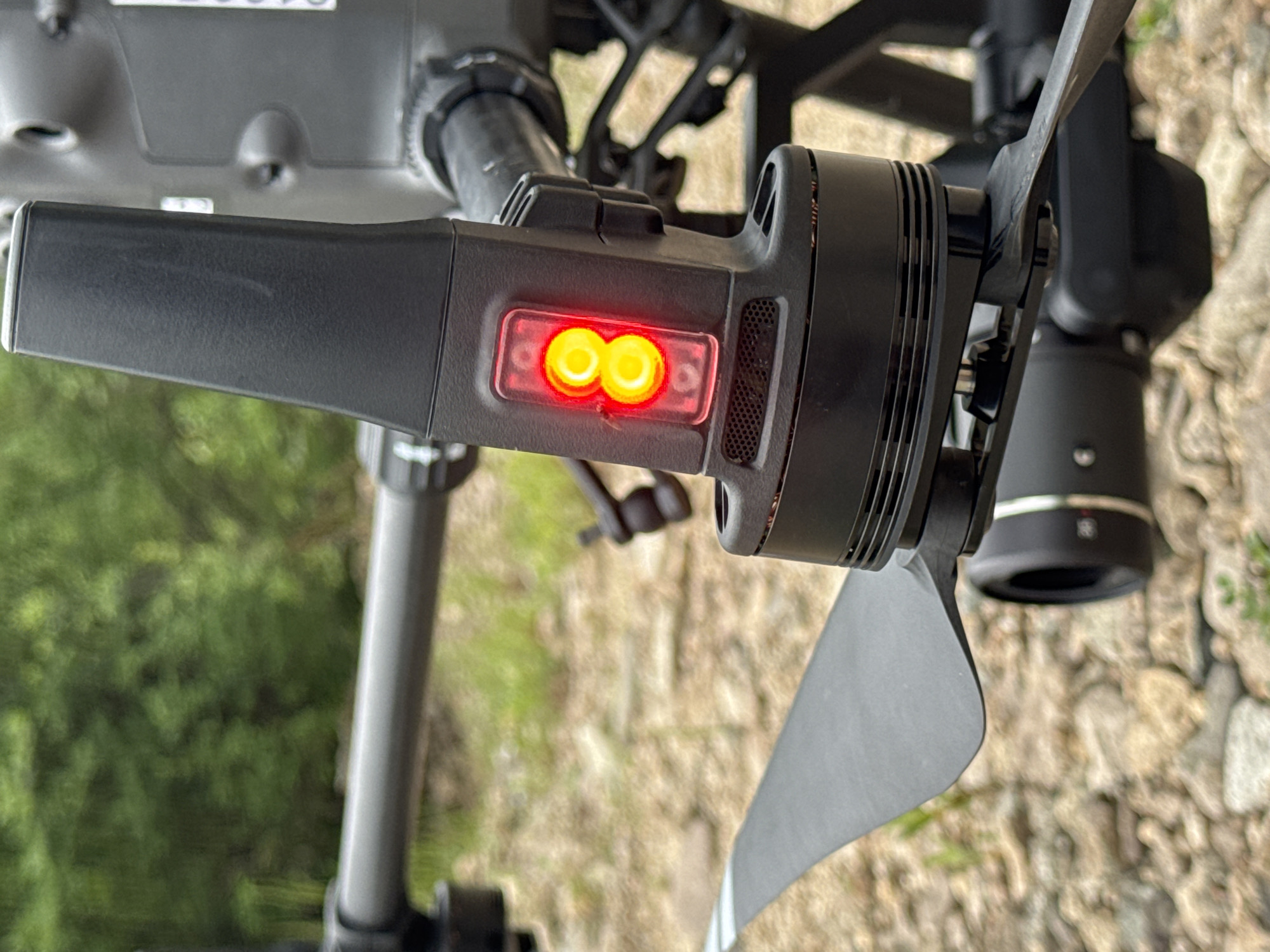 The image shows a close-up of a black drone with a red indicator light. The drone is on the ground, which is covered in small stones and patches of grass. In the background, there are blurred green trees. The drone appears to be a professional-grade model, possibly used for aerial photography or surveillance. The red light is on, suggesting the drone is active or preparing for flight. The focus is on the drone's arm and propeller. There are no people or animals visible in the image. The environment appears to be outdoors, possibly in a natural or semi-urban setting. The lighting suggests it is daytime. The location in Murtygit, Russia is not evident from the visual information in the image.
