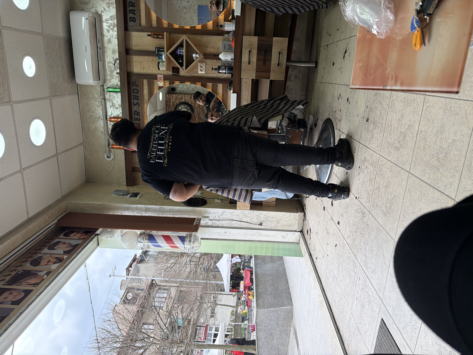 A view from inside a barbershop, the camera positioned near the entrance. The barber, wearing a black t-shirt that reads "Barbershop Deluxe" on the back, stands attending to a customer seated in a barber chair. The shop features wooden shelving stocked with various barbering products. Mirrors reflect the scene, showing the barber and customer from different angles. Through the glass storefront, a street scene in Panagyurishte, Bulgaria is visible, including buildings, trees, and a market with tents. The sky is overcast. A red and white barber pole stands outside the shop. The floor inside the shop is tiled. Hair clippings are visible on the floor around the barber chair.