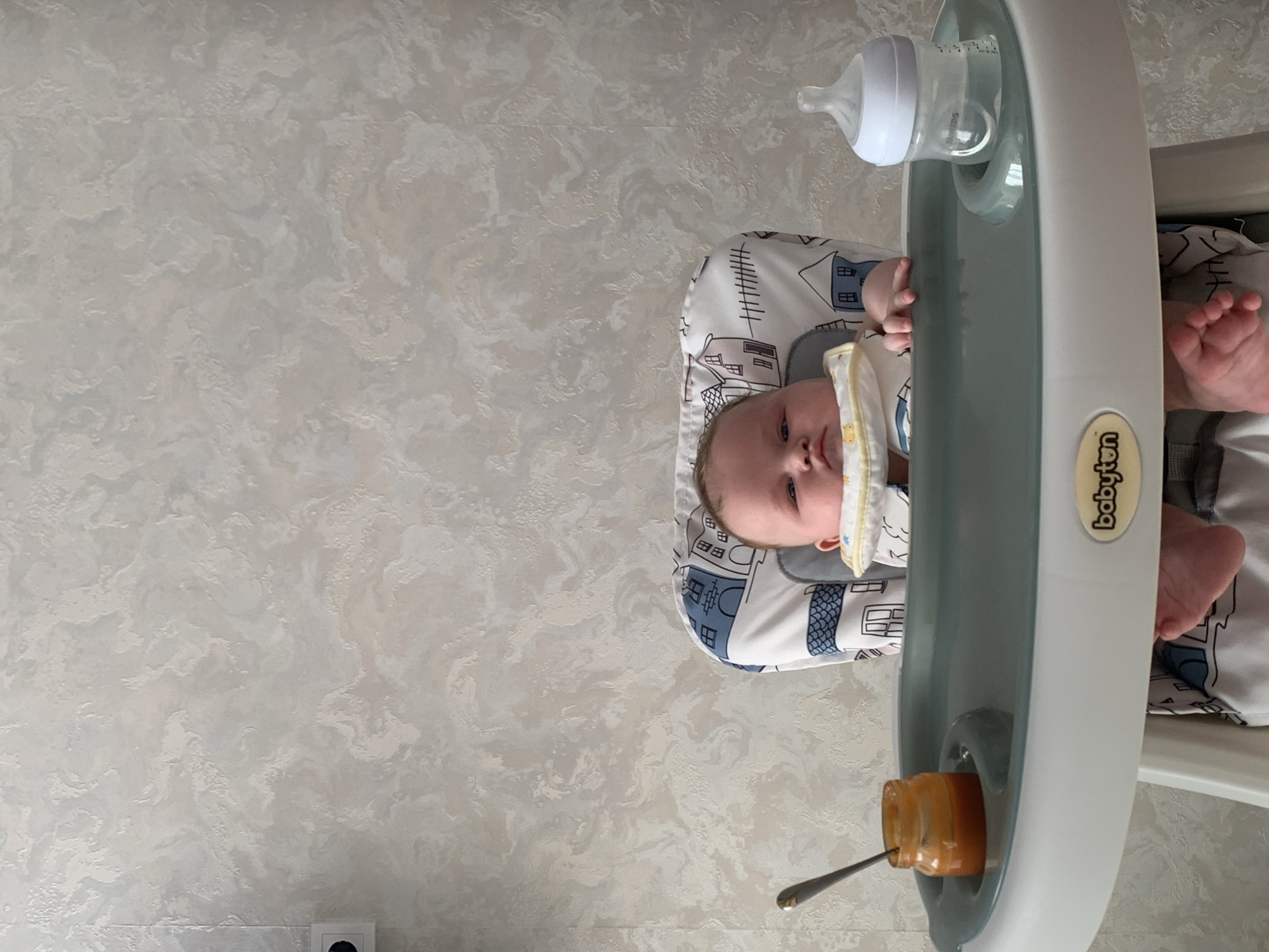This is a close-up, high-angle, portrait-oriented photograph of a baby in a high chair. The baby is facing forward, looking at the camera with a neutral expression. The baby is wearing a white onesie with blue and white houses printed on it and a yellow bib. The high chair has a grey tray with a bottle of water and a jar of baby food with a spoon in it. The background is a marble-patterned wall. The image appears to have been taken indoors, during the daytime. There is no information about the weather or specific location within Stavropol, Russia.
Visible text: "babyjem" is visible on the side of the high chair.