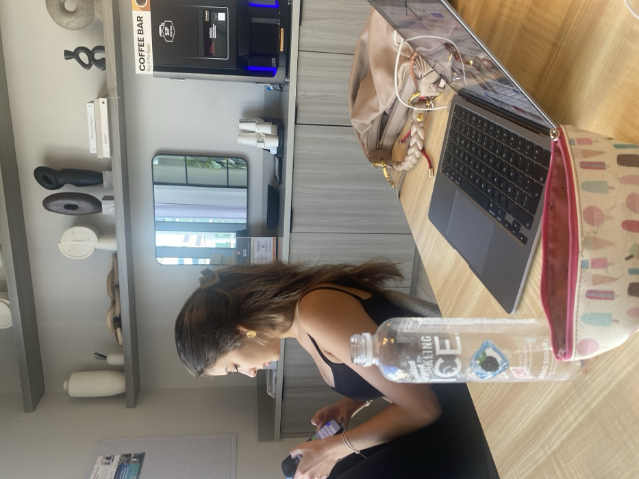 The image depicts an indoor common area or lounge within a modern building, consistent with the urban landscape of Miami, United States of America. A female subject is seated in profile on the left, possessing long dark brown hair secured with a visible clip. She wears a black tank top and a golden oval-shaped earring on her visible ear. A light-colored bracelet is present on her left wrist. She is actively looking down at and operating a black smartphone held in both hands.
In the foreground, on a light-colored wooden table, a clear plastic bottle of "Sparkling ICE" (Black Raspberry, Zero Sugar) stands upright. Immediately in front, a small, open pouch with a white background and a colorful popsicle pattern is visible. To the right of the bottle, an open silver-colored laptop is positioned, its screen angled upwards. Behind the laptop, a beige handbag with a braided handle and gold hardware rests on the table.
The background features a built-in counter with light-toned paneled cabinets below. Above the counter, two floating shelves display various decorative objects, including white ceramic vases, abstract sculptures in white and dark tones, and a stack of books. A rectangular mirror with a black frame, reflecting modern high-rise buildings and foliage, is centered above the counter. To the right of the mirror, a black "SIP" branded coffee machine, labeled "COFFEE BAR, Try it for free," is visible with blue illuminated accents and a stack of white disposable cups. A dark tray and a small informational sign displaying "20" are also present on the counter. On the far left, a white board with printed documents is partially visible.