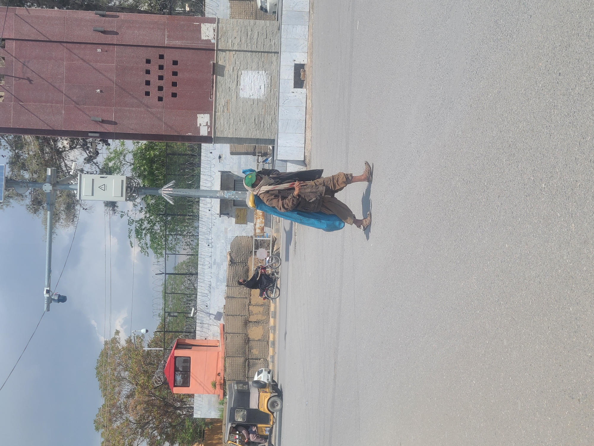 A man wearing a green cap and carrying a blue and yellow object walks along a street in Quetta City Tehsil, Pakistan. Barbed wire, stacked sandbags, and a small security post are visible along the side of the road. A motorcycle and a tuk-tuk are also present.