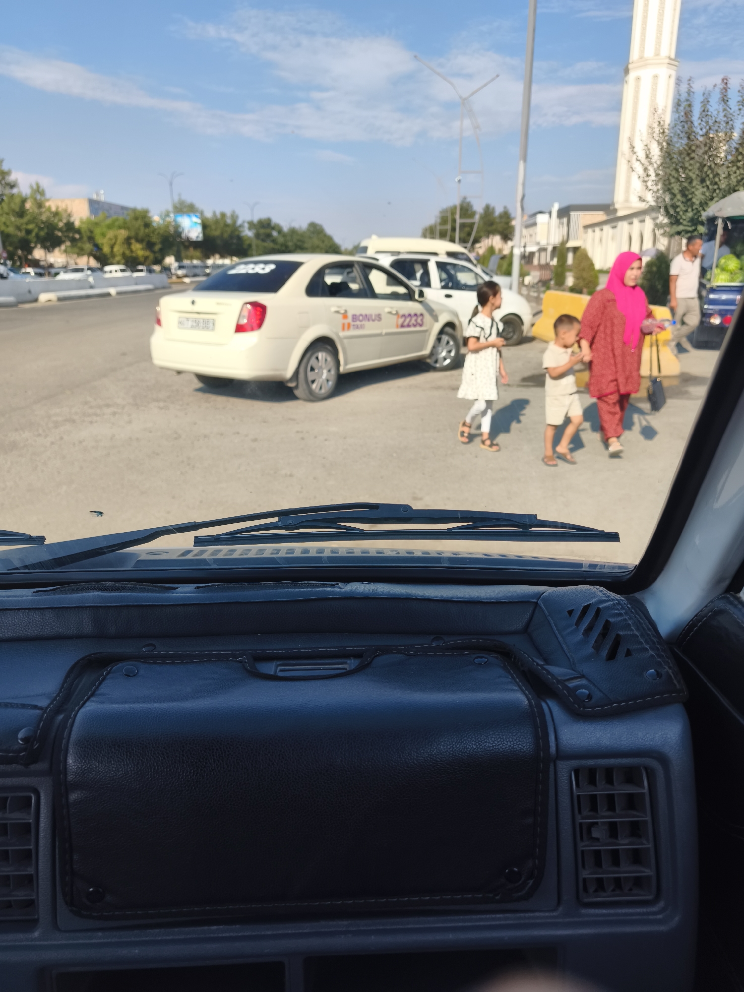 The image shows a street scene in the city of Buz, Uzbekistan, taken from inside a vehicle. In the foreground, the interior of the vehicle is visible, including the dashboard and windshield wipers. Outside the vehicle, a white taxi with "BONUS TAXI 2233" written on it is parked on the street. A woman in a pink hijab and a red outfit is walking with two children. The street is paved, and there are trees and buildings in the background. The sky is clear and blue.