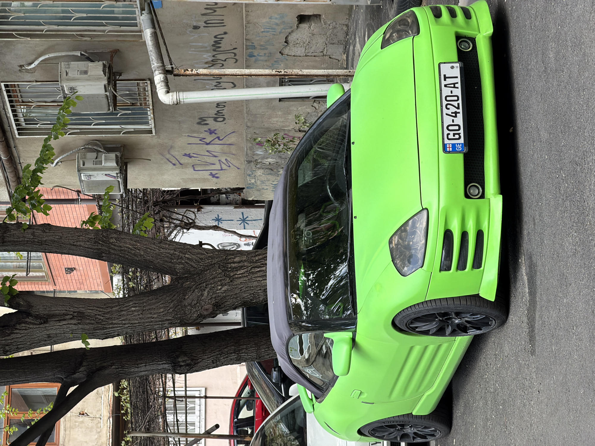 A vibrant, neon-green sporty car, possibly a convertible or coupe, is parked on a paved street in Tbilisi, Georgia. The vehicle features a custom front bumper with a black mesh grille and round fog lights. Its Georgian license plate, "GO-420-AT," is centrally mounted, displaying the "GE" country code. The car is equipped with black multi-spoke wheels and Michelin tires.
In the background, an old, dilapidated building with concrete walls is visible. Two antiquated, external air conditioning units are mounted on the wall, alongside windows protected by metal grilles and various utility pipes. The building's walls are heavily covered in graffiti, including script that appears to be Georgian, as well as abstract symbols and Roman letters.
A large tree with a thick, textured trunk and some green leaves stands prominently on the left side, partially obscuring parts of the building and other parked vehicles. Hints of a red car and a white car are visible behind the bright green vehicle. The scene is captured in daylight, suggesting clear weather conditions.