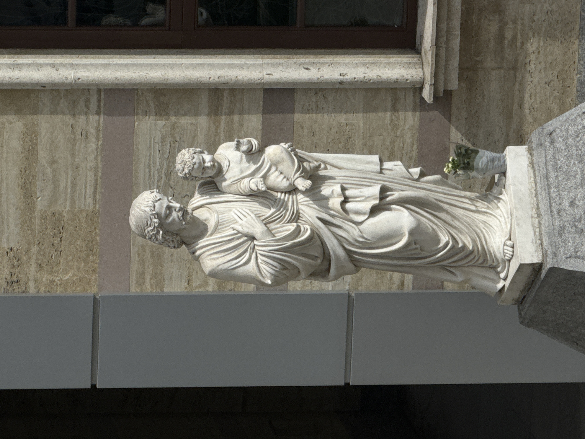 A white marble statue, centrally positioned, depicts an adult male figure identified as Saint Joseph holding a child figure identified as Jesus. Saint Joseph, characterized by a beard, long hair, and flowing robes, holds his right hand over his chest while supporting the Child Jesus with his left arm. He looks downwards towards the child. The Child Jesus, with curly hair and partial draping, extends his right hand with an open palm and holds a spherical object in his left hand, looking forward. The statue rests on a white rectangular base, which is set upon a larger, multi-tiered grey stone pedestal.
To the right of the statue's base, a small white pot containing green foliage and white flowers is placed. The entire setup is situated outdoors against the façade of the Co-Cathedral of St Joseph in Sofia, Bulgaria. The immediate background features a wall constructed from light brown, horizontally textured stone panels, with a darker brown horizontal strip running across the mid-section. To the left of the statue, a modern-style grey architectural column, composed of two stacked rectangular sections, partially obscures a dark recessed area. To the right, a vertical light-colored stone pilaster stands beside a dark brown multi-paned window. Below the window, a light-colored stone windowsill is visible. The scene is illuminated by direct sunlight originating from the left, casting distinct shadows on the building wall and from the architectural elements.
