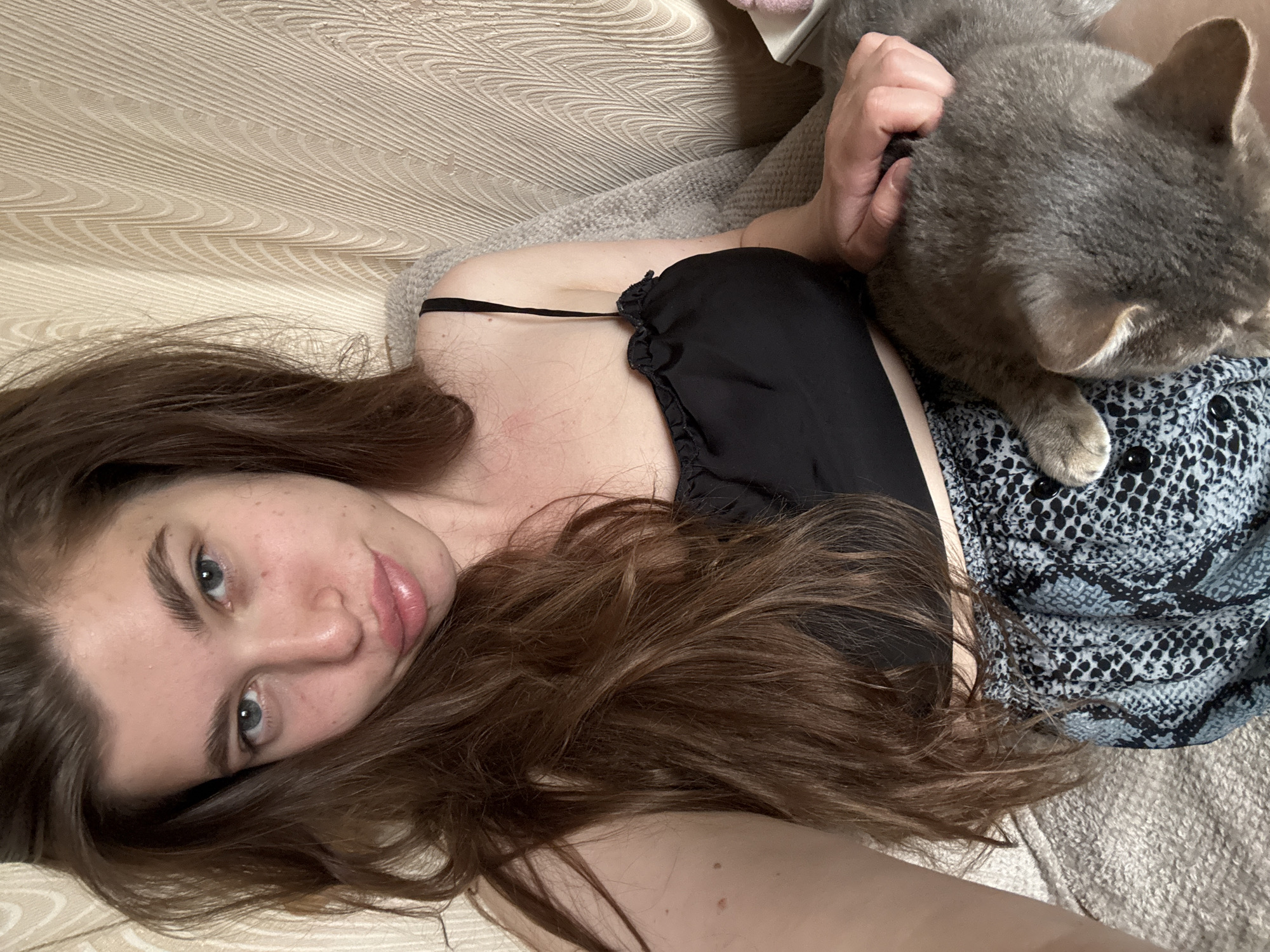 A woman with long brown hair and blue eyes is lying down on a couch or bed with a gray cat on her torso. The woman is wearing a black top and a patterned skirt or shorts. The cat is mostly gray. The location in the city of Мещёры, Russia cannot be determined from this image.
