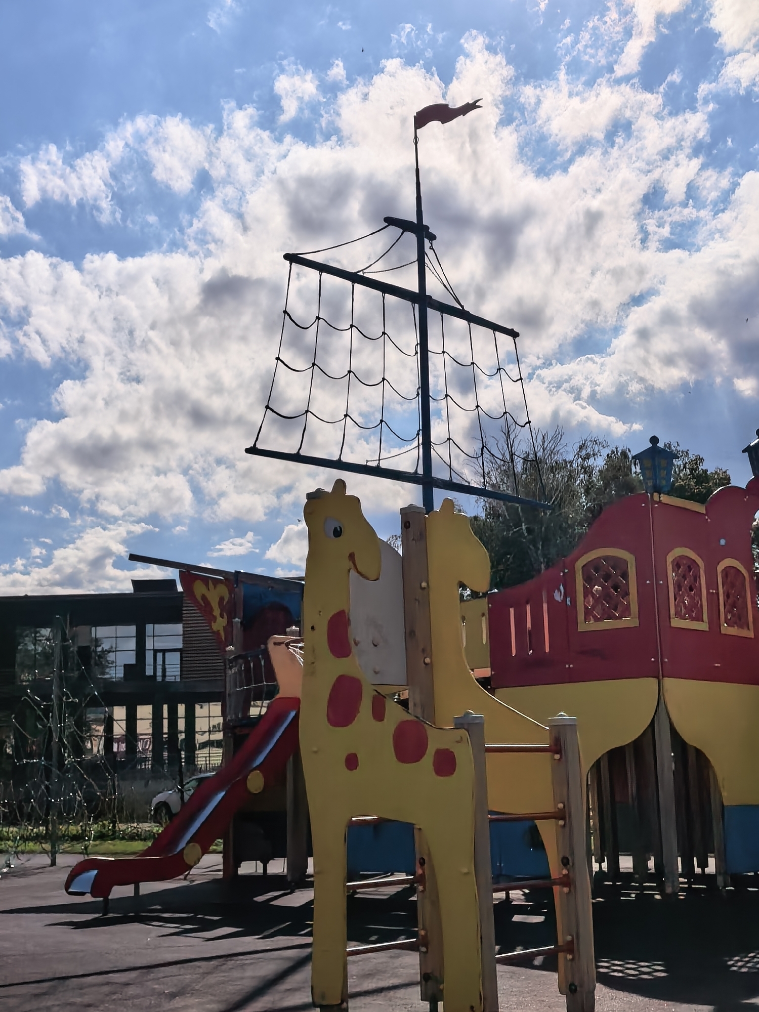 A colorful children's playground designed as a pirate ship sits under a partly cloudy sky. The ship has a prominent mast with netting resembling sails, topped with a red flag. In the foreground, a yellow giraffe-shaped structure with red spots is part of the play equipment, along with a red slide. Behind the ship, a modern building with large windows is visible, suggesting the playground is in a developed area. Trees are also present in the background. The overall impression is a bright, playful environment for children. The time of day appears to be daytime, with sunlight casting shadows. The weather seems pleasant, with a mix of sun and clouds. The location is Sterlitamak, Russia. There is no visible text in the image.