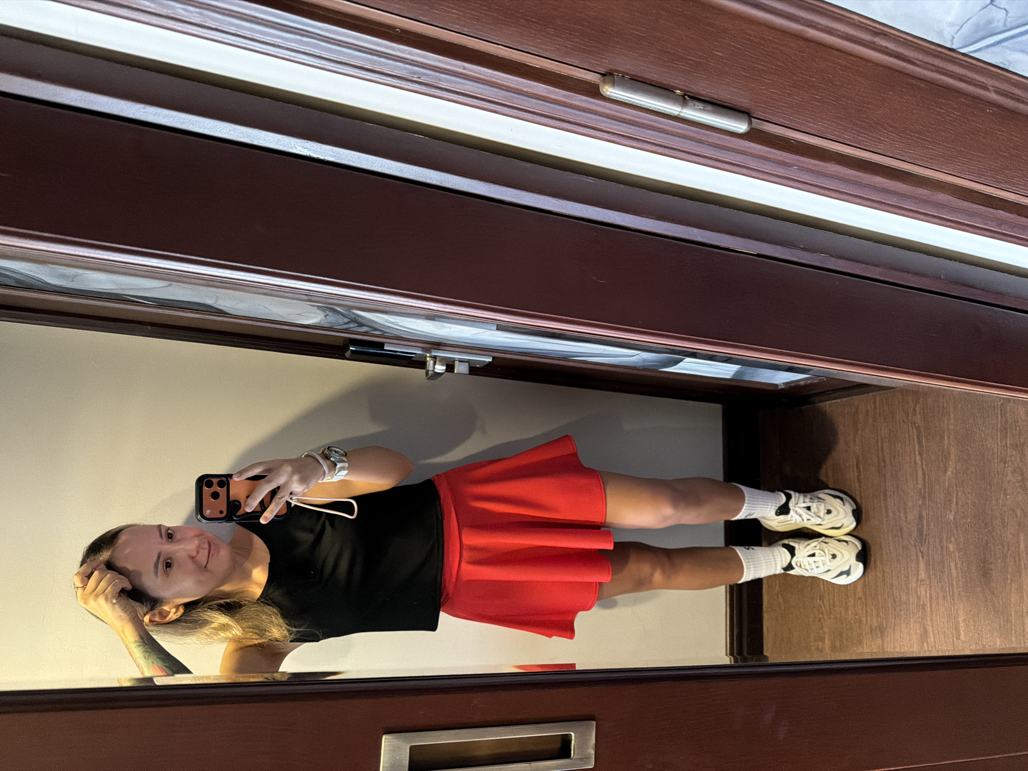 A woman is taking a selfie in a room, reflected in a large mirror. She is wearing a black tank top, a red mini skirt, white socks, and white athletic shoes. Her hair is blonde and pulled back, and she is wearing a watch on her left wrist. She is holding her phone in her right hand and touching her hair with her left hand. The background consists of a plain wall and a wooden door frame. There are no visible elements that suggest the location is Thuận Hóa Ward, Vietnam.