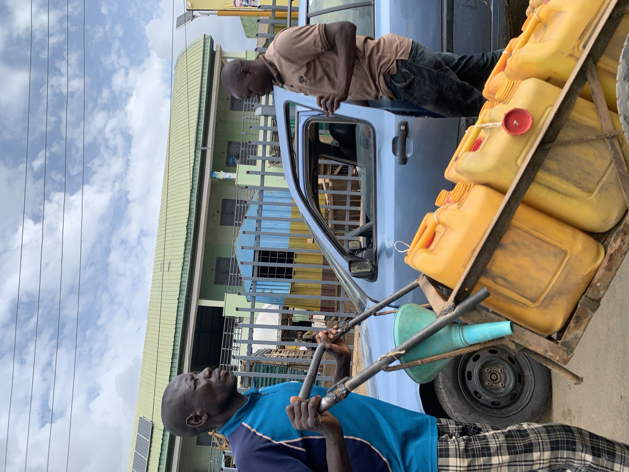 In Akwanga, Nigeria, two African men are present next to a light blue car. One man, wearing a brown shirt and dark pants, is leaning into the car through the open driver's side door. The other man, wearing a blue shirt and checkered pants, is pulling a metal cart loaded with yellow jerrycans. The background includes a building with a green and white facade and a blue and yellow fence. The sky is cloudy.