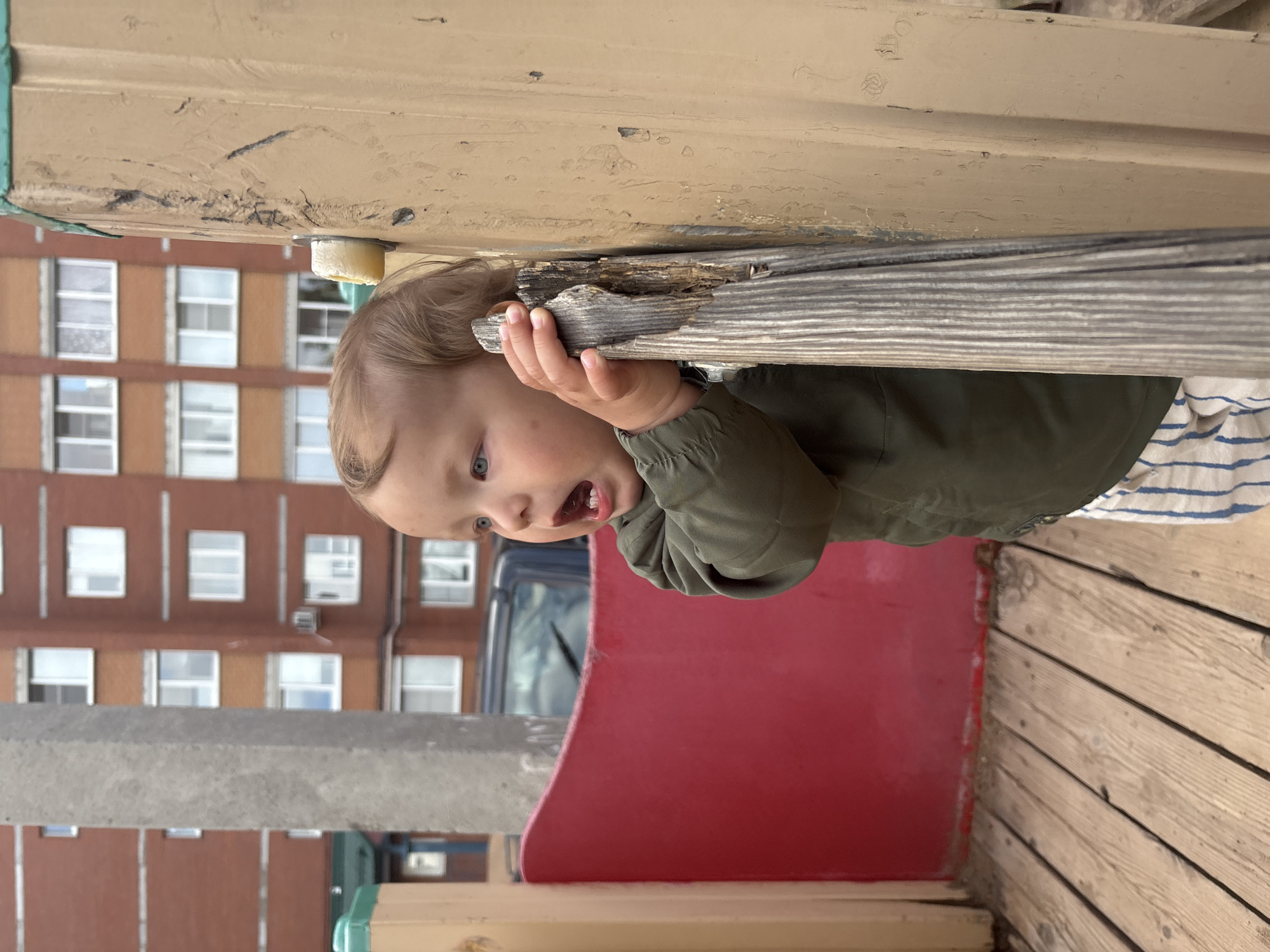 A toddler with blonde hair and blue eyes, wearing a green jacket and striped pants, stands outdoors, holding onto a weathered wooden post. The child is looking to the left, with their mouth open as if speaking or surprised. Behind the toddler, a bright red curved wall is visible, and beyond that, a multi-story brick building with numerous windows dominates the background. The foreground consists of a wooden platform with planks running diagonally. The scene appears to be a playground in a residential area of Chaikovsky, Russia.