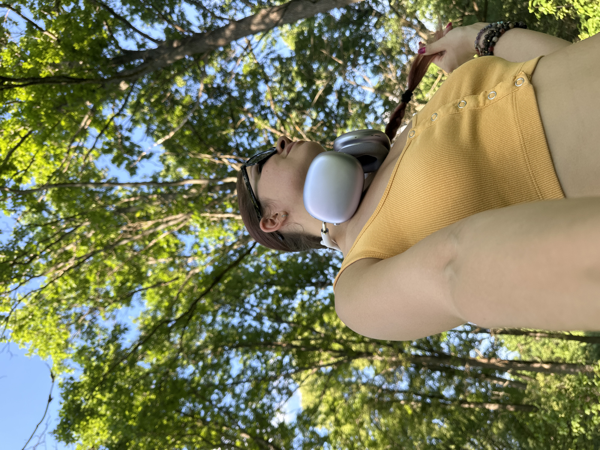 In this low-angle, outdoor shot, a young woman with brown hair styled in a braid is seen from the chest up, looking to her right and upwards. She is wearing a yellow cropped tank top with silver buttons and large silver headphones around her neck. Her left arm is extended towards the camera, and she is touching her braid with her right hand. She is also wearing sunglasses and bracelets on her right wrist. The background is filled with lush green trees and a clear blue sky, suggesting a park or wooded area, possibly within the city of Vladimir, Russia, during daylight hours.
