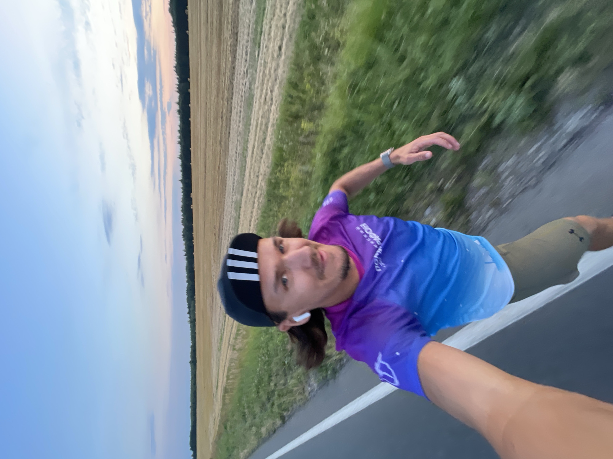 A selfie-style, waist-up shot shows a man with shoulder-length brown hair, wearing a black baseball cap with white stripes, white wireless earbuds, and a purple and blue gradient t-shirt with white lettering. He is also wearing olive-green shorts and a wristwatch. The man is in motion, likely running or cycling, as indicated by the blurriness of the background and the slight motion blur on his arm and hair.
He is positioned on the left side of a paved road, which has a white line marking the edge. To the right of the road is a strip of green grass and then a large, golden field that appears to be harvested, with rows of stubble visible. Beyond the field, a dark line of trees forms the horizon, and above that, a pale blue sky with some wispy clouds transitions into a softer hue near the horizon, suggesting it is either dawn or dusk. The overall environment is rural, with fields and a road. The specific location in the city of Ol'khovo, Russia, is not explicitly indicated by any visual cues in the image itself, but is provided as context.