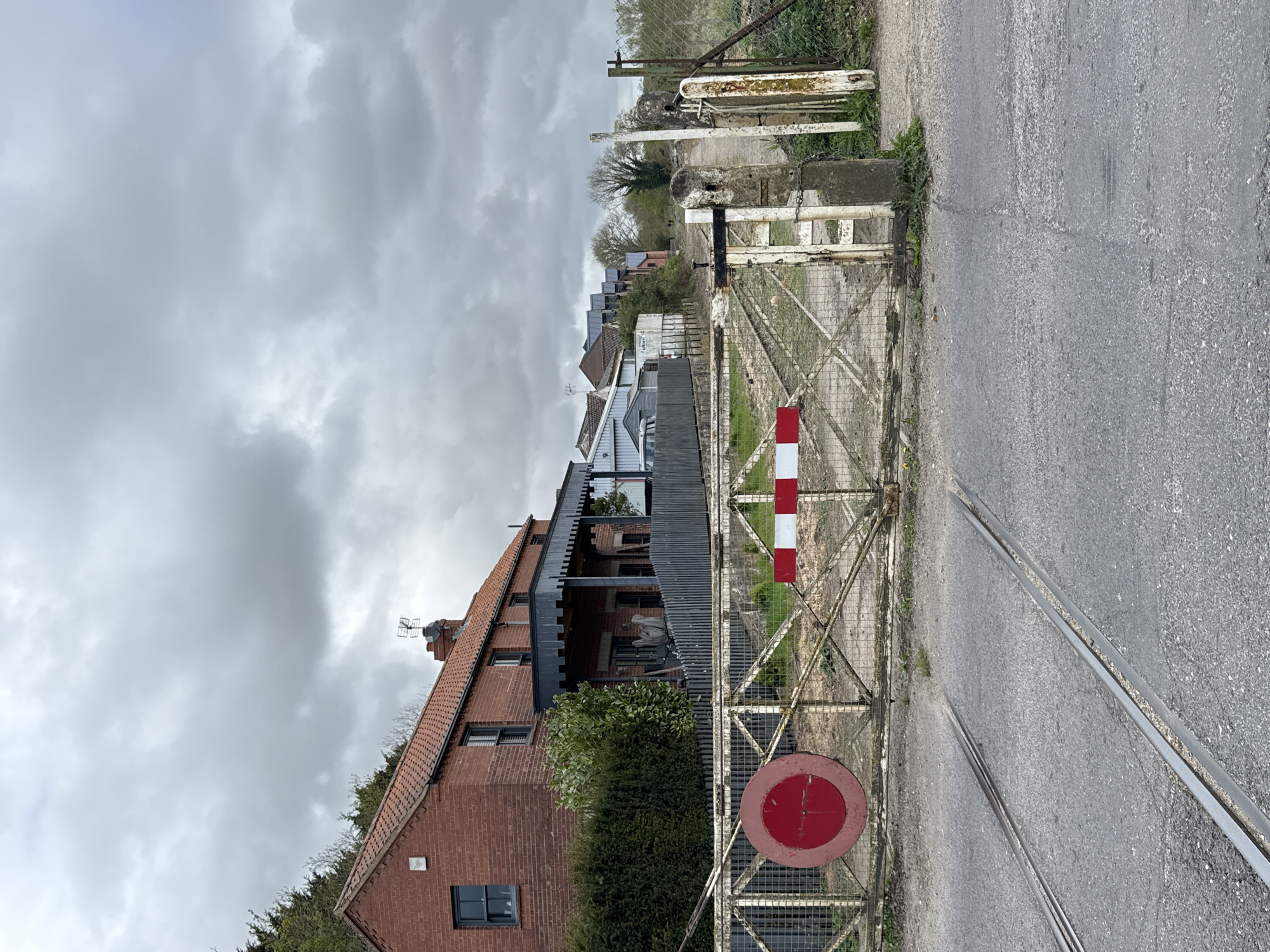 The image depicts a static outdoor scene, likely a rural railway crossing or a disused railway line in the Breckland District, United Kingdom, under an overcast sky. In the foreground, a weathered metal wire-mesh gate stretches across the frame, featuring a red and white striped barrier arm and a red circular sign with a white horizontal bar, resembling a "no entry" or "road closed" sign. Faint, illegible text is visible on the white bar. Two railway tracks are embedded within the paved ground, running from the bottom right towards the gate.
To the left of the gate, a prominent brick house with a tiled roof, a visible chimney, and a television aerial stands, partially enveloped by green hedges and bushes. Behind this, other residential buildings and structures are scattered along the railway line, interspersed with trees and additional fencing. The sky above is entirely covered by dense, grey clouds, indicating an overcast daytime. There are no people visible, and no specific activities are taking place, contributing to a quiet, somewhat still atmosphere. The aged appearance of the gate and surrounding infrastructure suggests a long-established or potentially disused line.