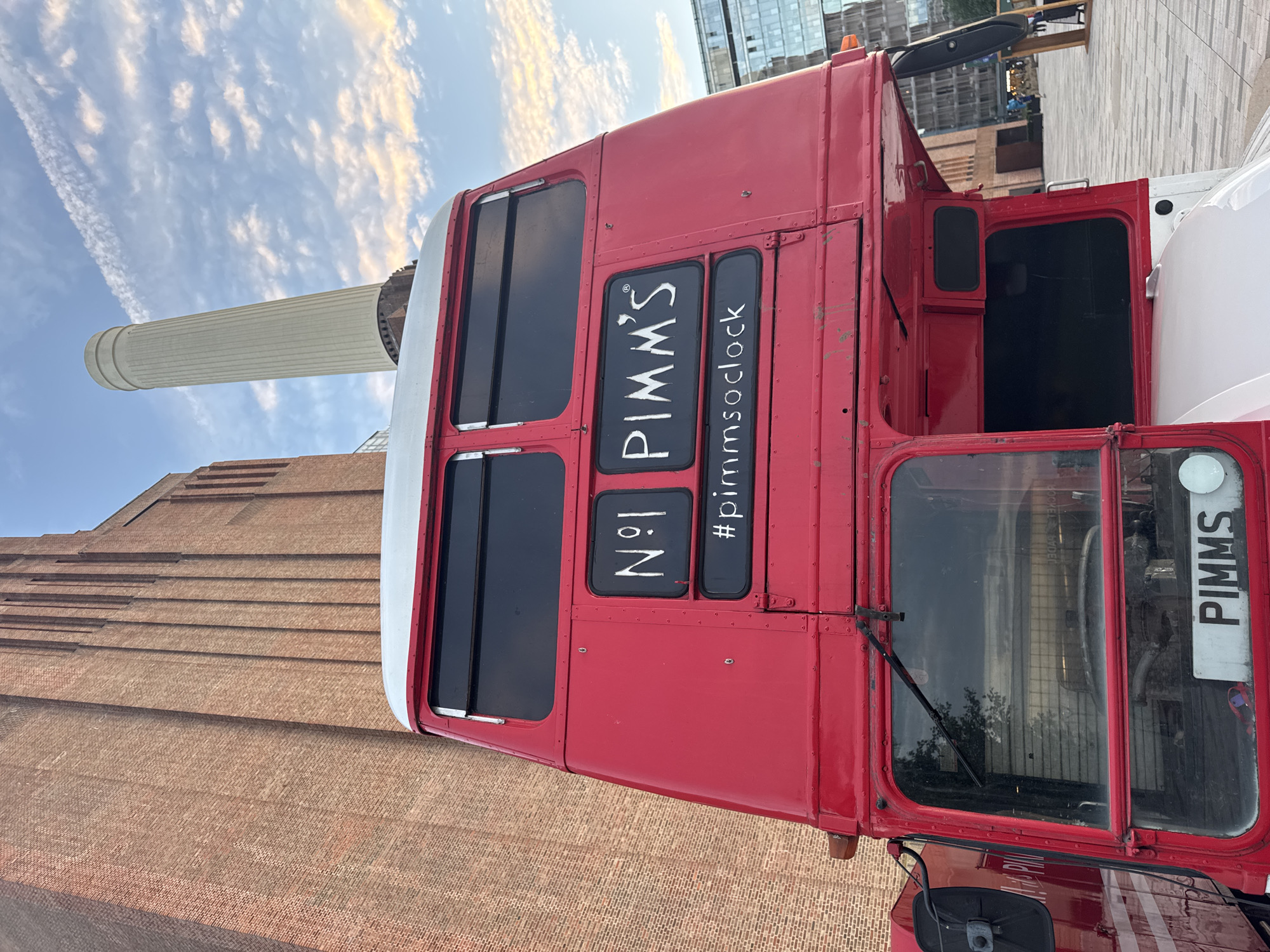 The image shows a red double-decker bus with "N°1 PIMM'S" and "#pimmsoclock" written on the side, parked in front of Battersea Power Station in London, United Kingdom. The power station, a Grade II listed building, is a prominent landmark. The sky is cloudy, with some contrails visible. The bus appears to be part of an event or promotion. The ground in front of the bus is paved with geometric patterns. The overall scene suggests an outdoor public space with a focus on the iconic London landmark and the branded bus.