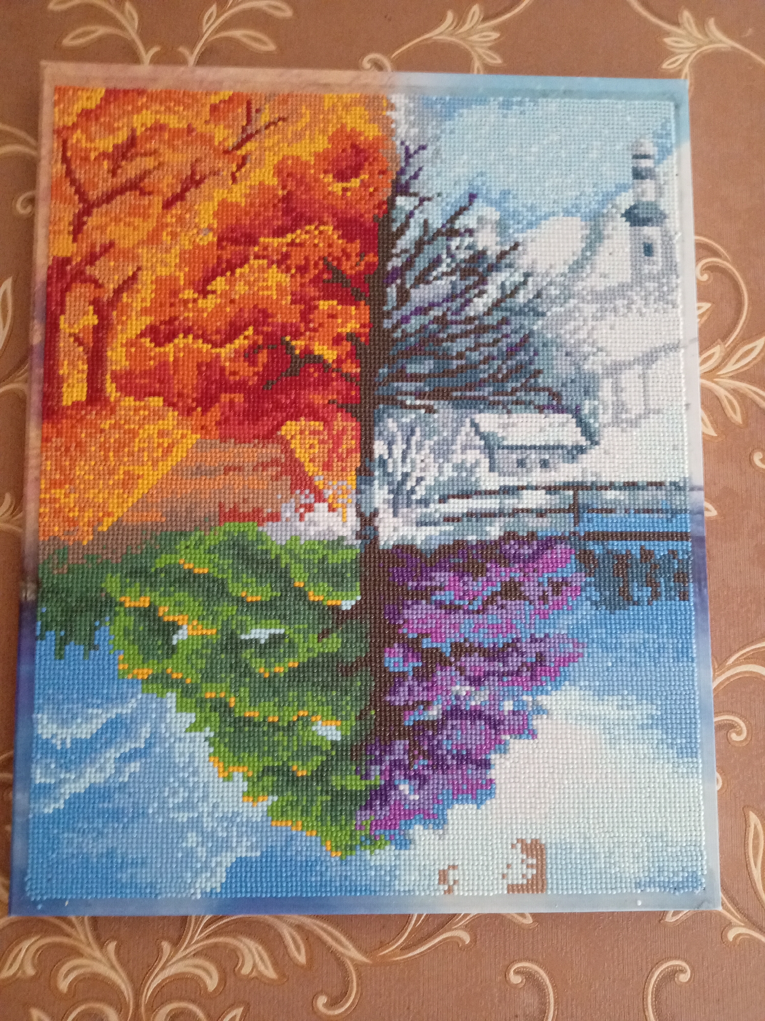 The image displays a four-panel diamond painting depicting the four seasons. The top left panel shows a tree with vibrant orange and red leaves, representing autumn. The top right panel portrays a winter scene with a snow-covered landscape and a lighthouse. The bottom left panel features a lush green tree, signifying spring, with a mountainous backdrop. The bottom right panel depicts a tree with purple flowers, suggesting summer, alongside a bridge. The painting is laid out on a patterned brown surface, likely a table or tablecloth. The diamond painting is framed with a clear plastic border. There are no people or animals present in the image, and the scene is not identifiable as being located in Kraskovo, Russia.