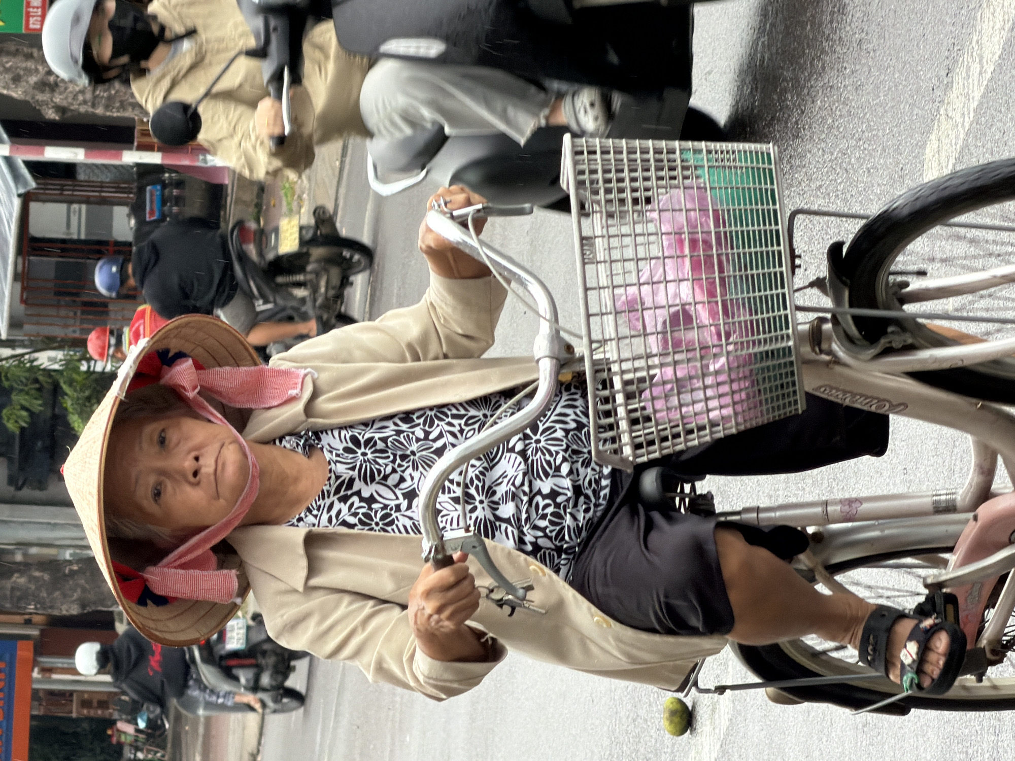 An elderly Vietnamese woman, wearing a traditional conical hat and a beige coat over a black and white floral shirt, rides a bicycle down a wet street in Nha Trang, Vietnam. She is holding the handlebars, and keys are visible hanging from them. Her bicycle has a wire basket in front containing pink and green plastic bags. To her right, a person on a scooter, wearing a tan jacket and a grey helmet, is also visible. In the background, other people on scooters and a person in a black hoodie on a scooter are also present. The street is wet, indicating it may be raining, and the environment suggests an urban setting with buildings and foliage.