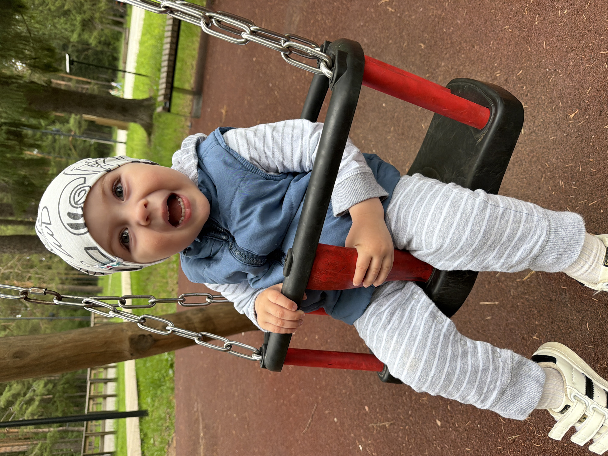 A young child wearing a white and black patterned beanie, a blue vest over a striped long-sleeved shirt, and striped gray pants is seated on a black swing seat with red supports. The child's hands are gripping the black bar of the swing. White sneakers with black stripes are on the child's feet. The swing is suspended by metal chains from a wooden structure. The background shows a playground with a reddish-brown rubberized surface, grass, trees, a wooden fence, and a park bench. The scene appears to be located in Votkinsk, Russia, likely in a public park or playground. The child's mouth is open, and they appear to be smiling or laughing.