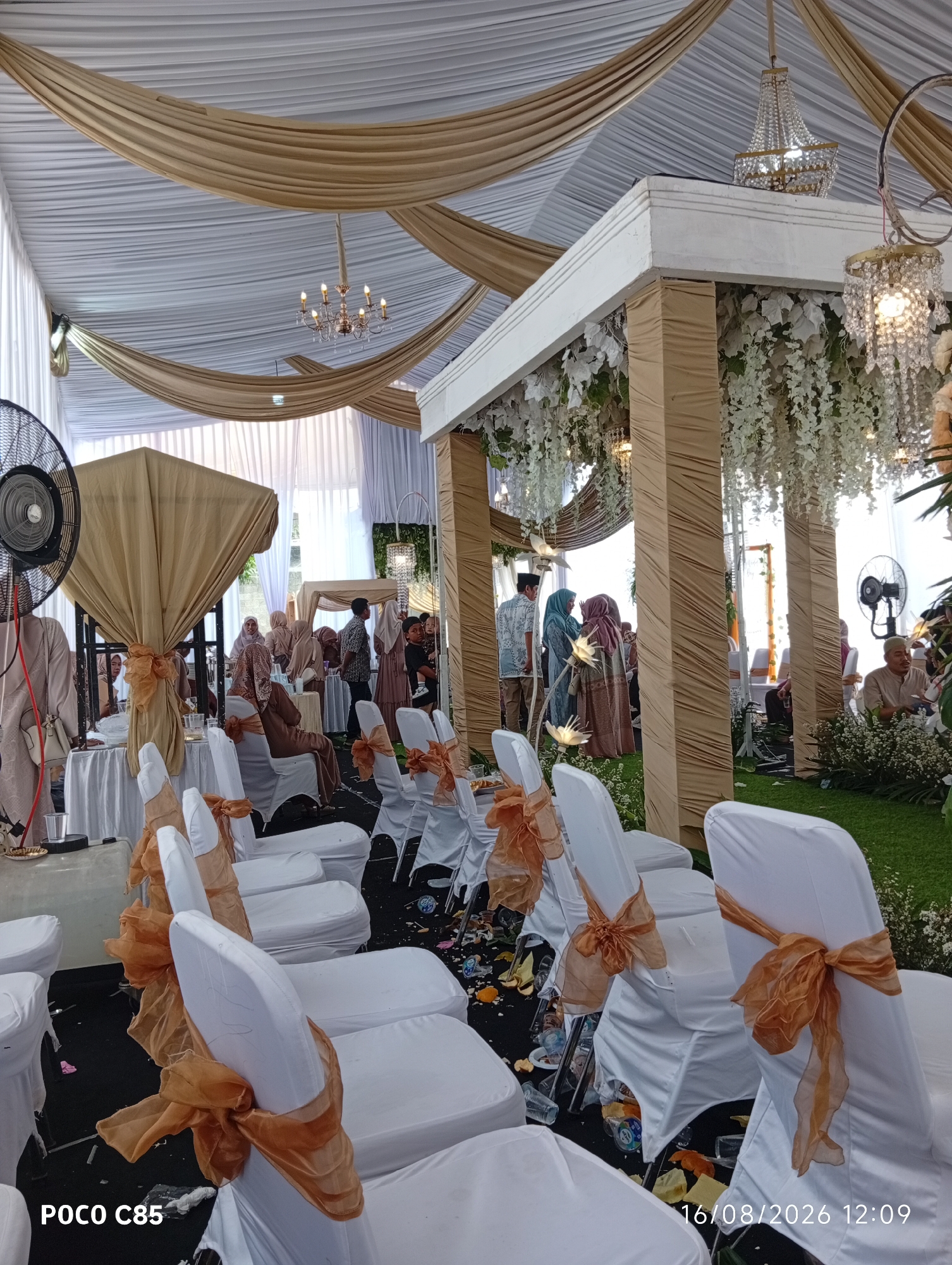 The image depicts an outdoor wedding reception or ceremony, likely in South Tangerang, Indonesia, given the presence of the "POCO C85" watermark which is a mobile phone brand. The scene is set under a large tent with white and gold draped fabric as a canopy, adorned with chandeliers. In the foreground, white chairs covered in white fabric with gold sashes are arranged on a black floor. Beyond the chairs, a grassy area is visible, with more white chairs and a ceremonial arch draped in white flowers and gold fabric. Several people are present, some seated and others standing, dressed in traditional Muslim attire. A large fan is visible on the left side of the image. The overall ambiance suggests a formal event with a festive and elegant decor.