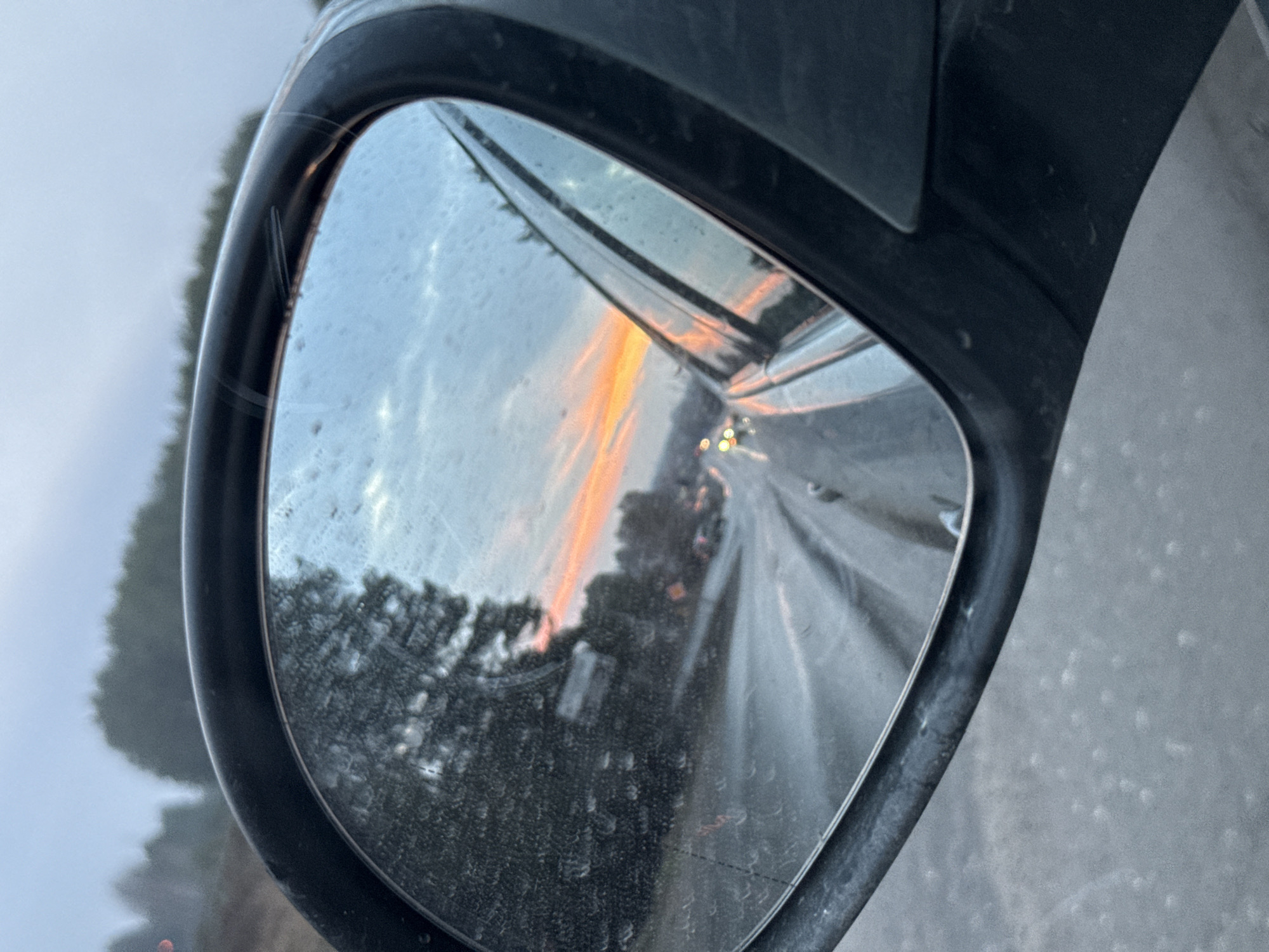 The image shows a car's side mirror reflecting a highway at sunset. In the reflection, multiple lanes of traffic can be seen, along with trees and a colorful sky reflecting orange and yellow hues. The mirror has water droplets. The photo appears to be taken on a cloudy day, and the location is not discernible as Chaikovsky, Russia.
