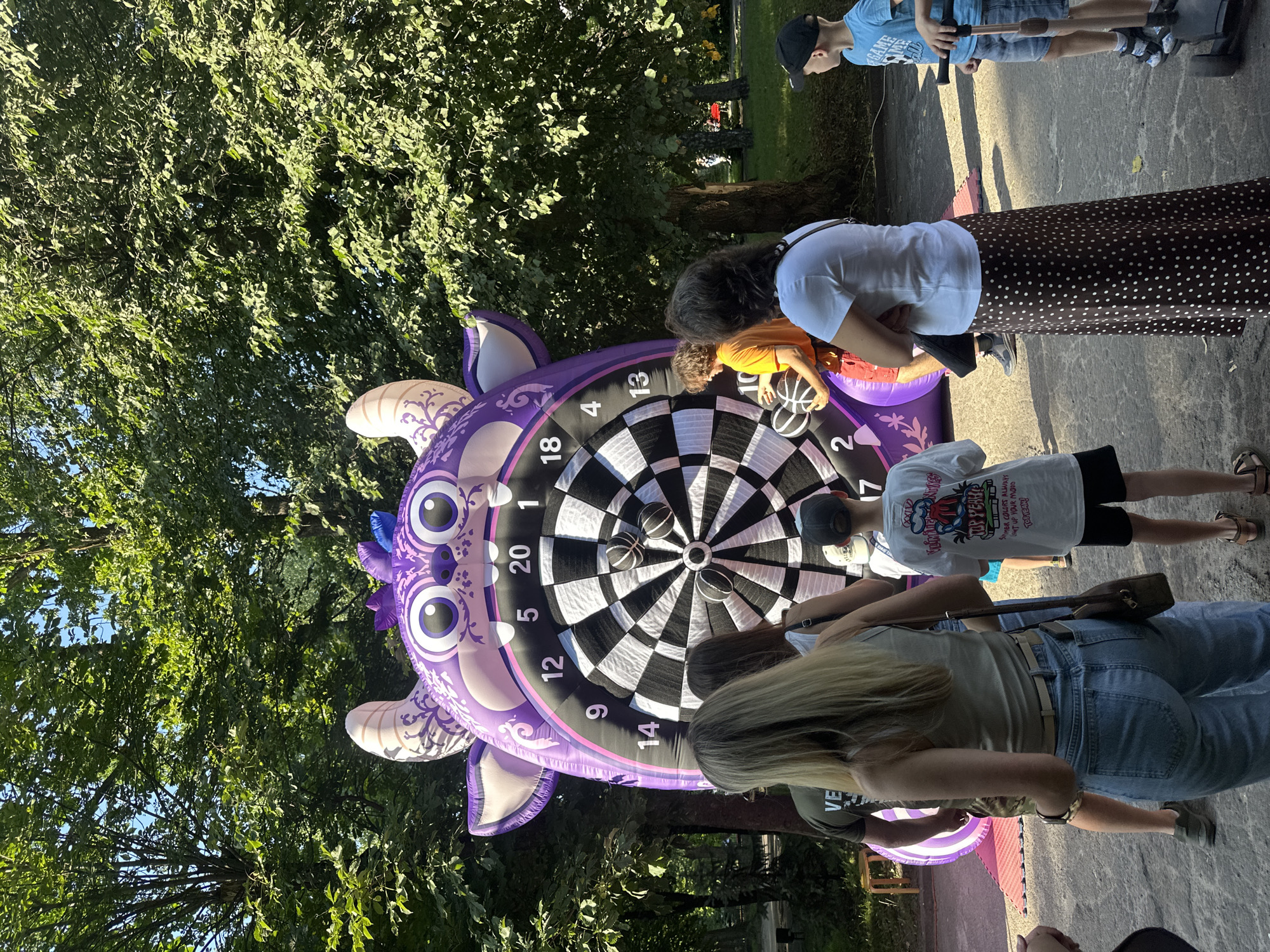 In the city of Ternopil, Ukraine, on a sunny day, a group of people gather around a large, inflatable dartboard shaped like a purple monster. The dartboard has numbers from 1 to 20, and several black and white balls are stuck in it. Some people are watching the game, while others are participating. A young boy in a blue t-shirt and shorts is standing to the right, looking towards the dartboard. Another boy in a white t-shirt is facing away from the camera, with his back to the dartboard. A woman in a white t-shirt and a brown polka-dot skirt is standing with her arms crossed, observing the game. In the background, lush green trees provide shade, suggesting a park or outdoor recreational area. The ground is a mix of dirt and paved surfaces.