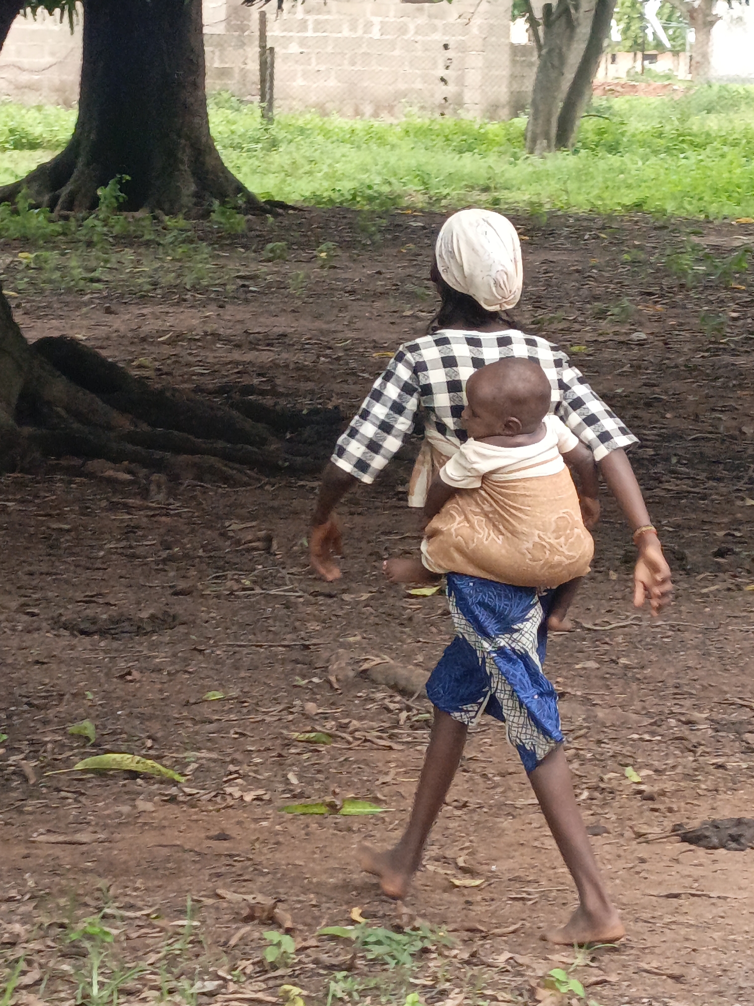 A woman wearing a white headscarf and a checkered shirt carries a baby on her back in a cloth wrap. She is walking outdoors on a dirt path, with a large tree on the left and green foliage in the background. She is wearing a blue and white patterned wrap around her lower body. The baby is facing away from the viewer and wearing a light-colored onesie.