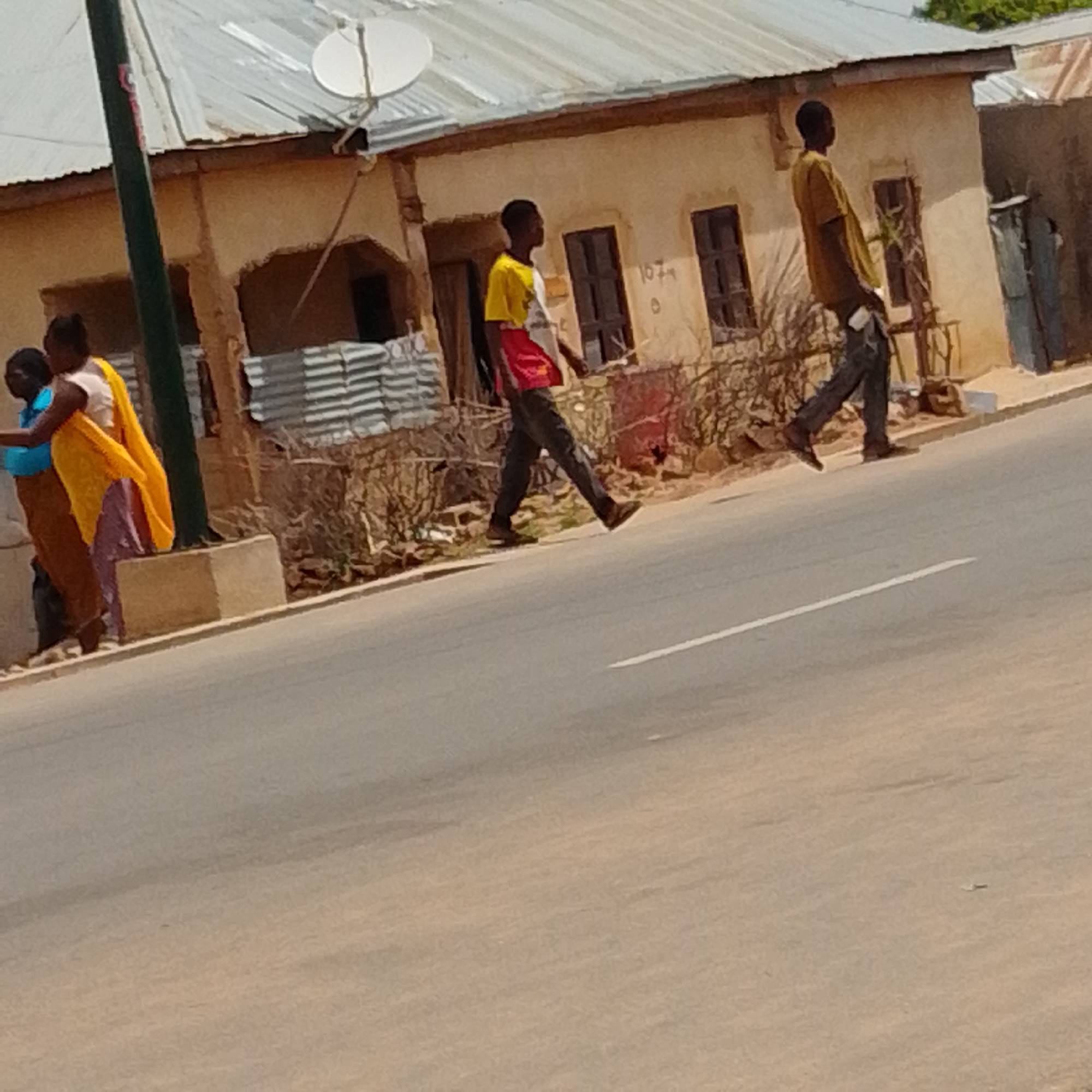 In Gombe, Nigeria, on a paved road, two young men are walking on a crosswalk. The man on the left wears a yellow and white t-shirt with red accents and dark pants. The man on the right wears a yellow t-shirt and dark pants. In the background, a light-colored building with a corrugated metal roof stands. A satellite dish is visible on the roof. To the left, three women are standing near a pole and a concrete structure. One woman wears a blue top, another a light-colored top with a yellow and purple patterned wrap, and a third is mostly obscured, wearing a brown garment. The ground is covered in asphalt, with a white line marking the crosswalk.