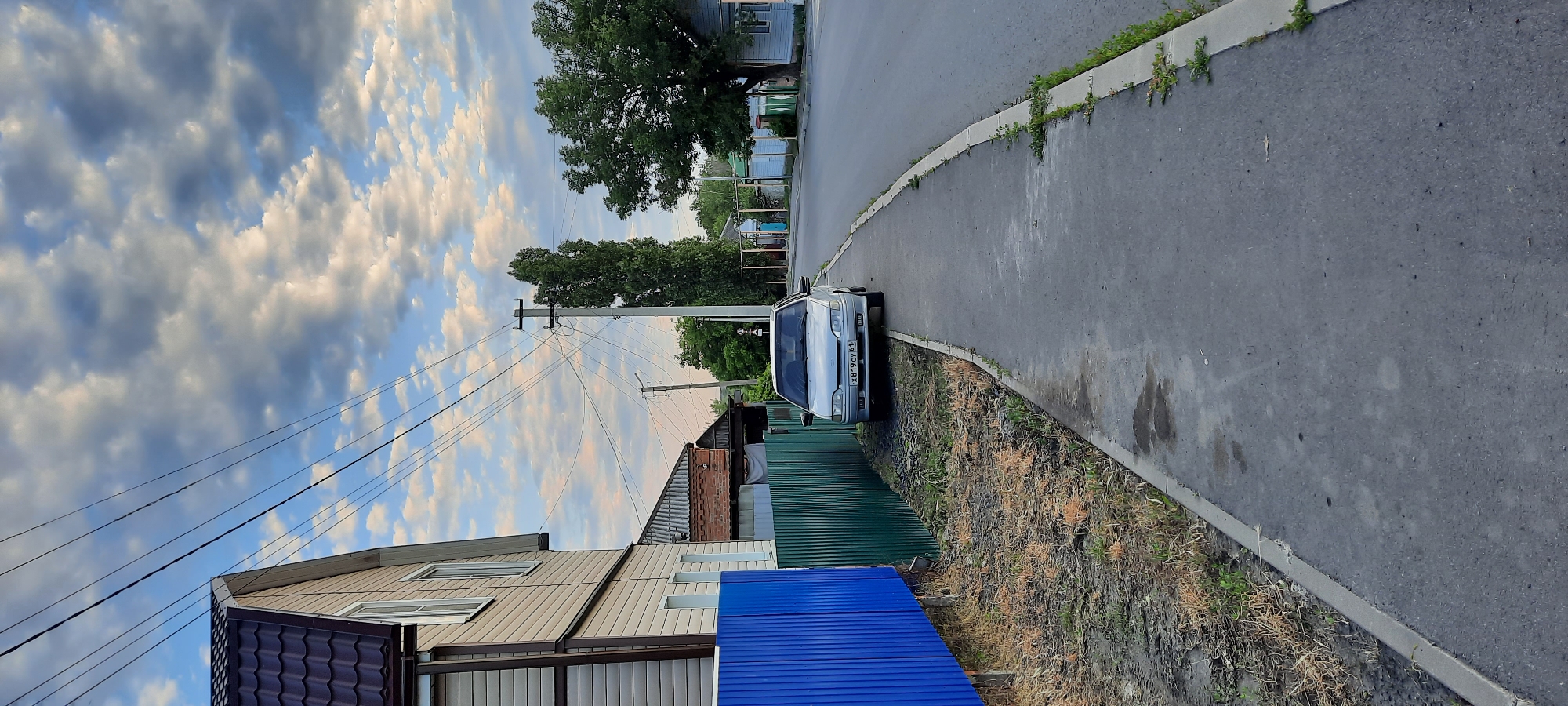 A light-colored sedan is parked on the side of a paved road in Shakhty City District, Russia. The car is positioned on the shoulder of the road, next to a sidewalk. A blue and green metal fence runs along the property line behind the car. To the left of the fence is a two-story house with beige siding and a dark brown roof. In the background, power lines crisscross the sky above a residential street lined with trees and other houses. The sky is partly cloudy with scattered white and gray clouds against a blue background. The scene appears to be a quiet, daytime residential setting.