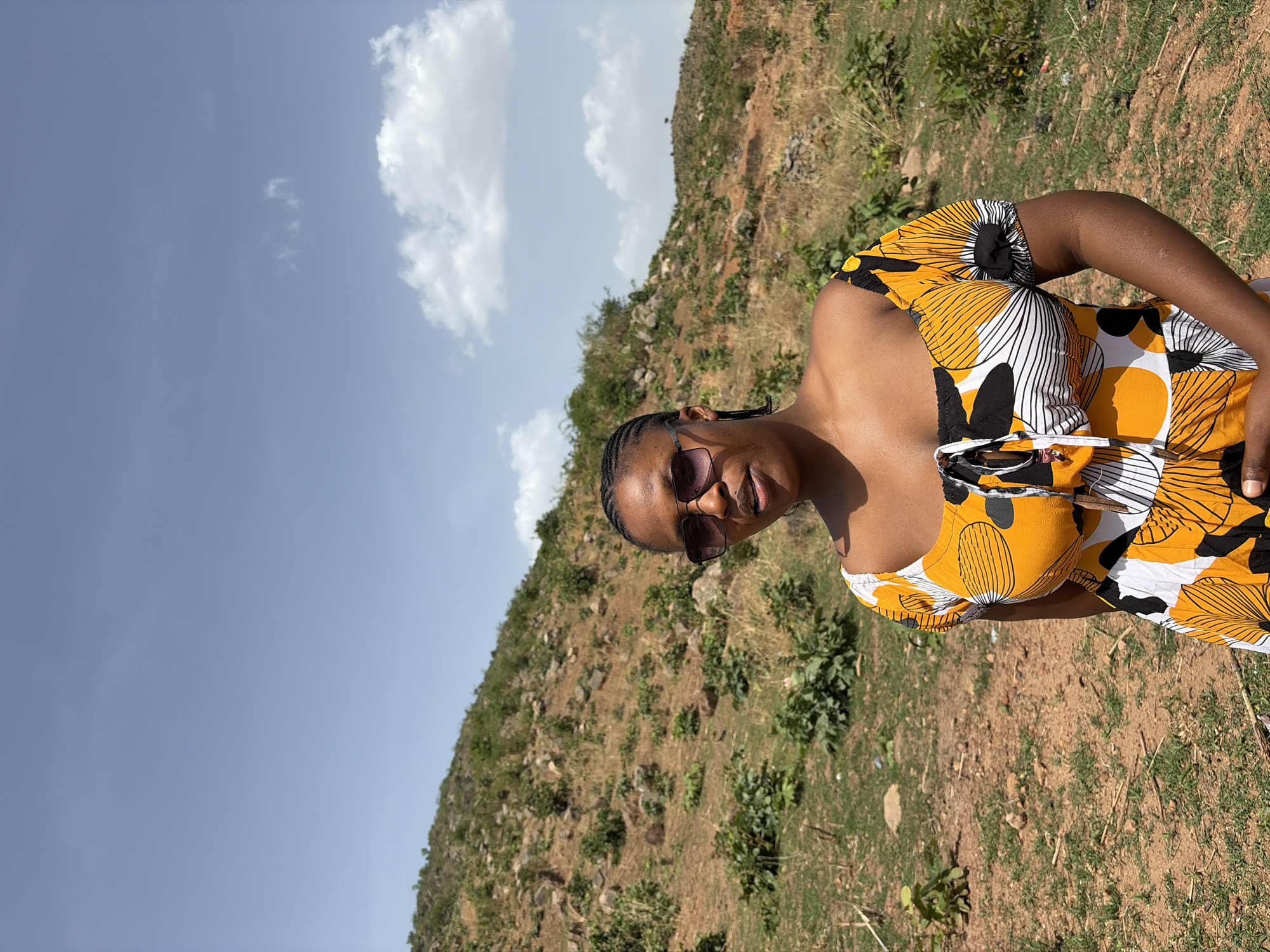 The image depicts a young Black woman captured in an outdoor portrait, rotated to appear vertical. She is shown from the waist up, looking towards the viewer with a subtle smile, wearing dark sunglasses that obscure her eyes. Her hair is styled in neat braids. She is dressed in a vibrant, short-sleeved or sleeveless dress featuring a bold pattern of yellow, black, and white abstract floral or starburst designs. Her skin tone appears warm, illuminated by bright sunlight.
The setting is a natural, hilly landscape. Behind her, a sloping hill is covered with sparse green vegetation, dry brownish grass, and exposed reddish-brown earth and rocks, suggesting a somewhat rugged and semi-arid terrain. Above the landscape, the sky is clear and bright blue with a few scattered white, fluffy clouds. The strong, direct sunlight indicates it is daytime, likely late morning or early afternoon.
The woman appears relaxed, posing for the photograph in this scenic environment. No visible text is present in the image. Given the contextual information, the location is Jos, Nigeria, which aligns with the described landscape and sunny weather.