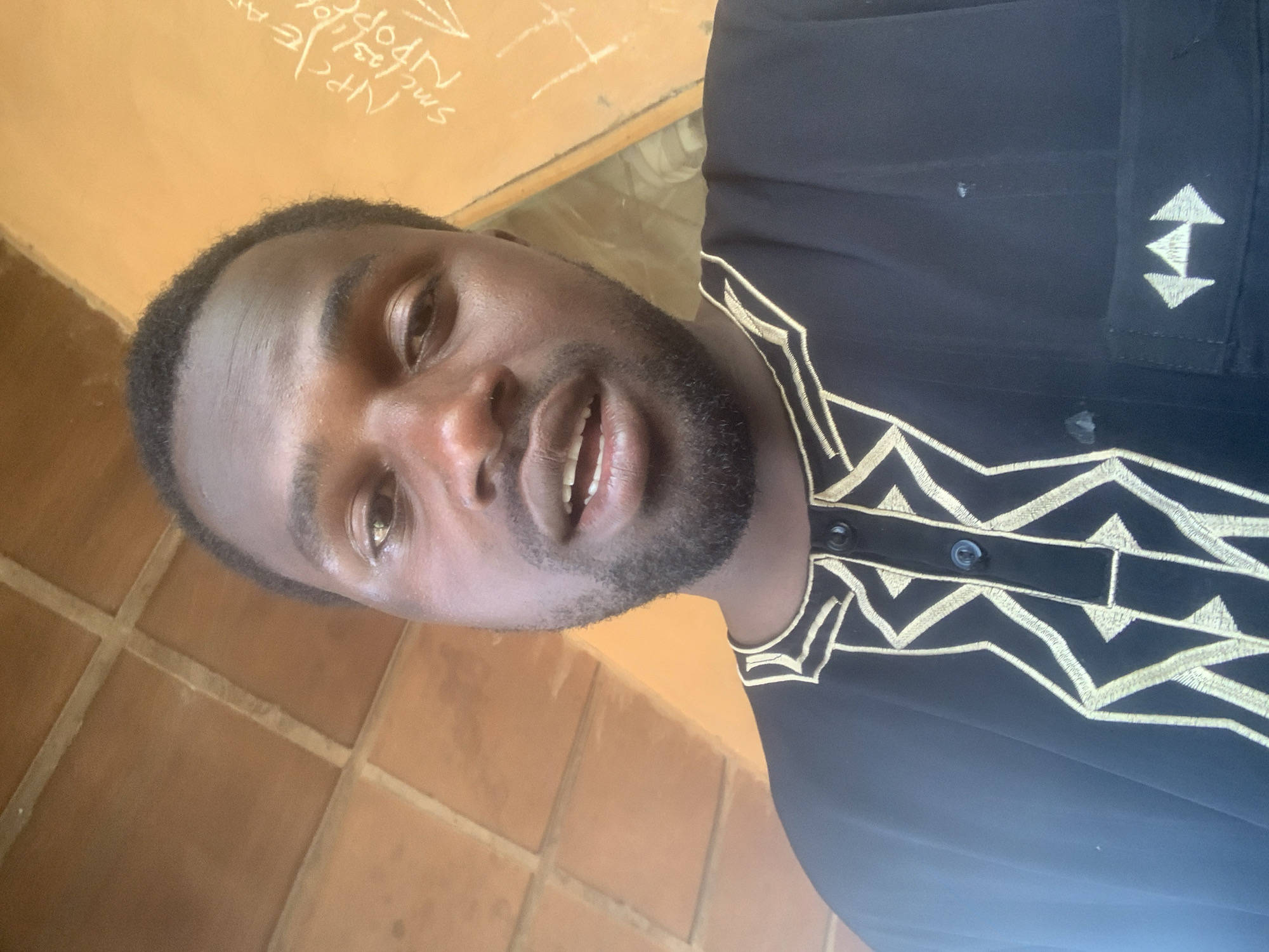 This media file shows a close-up, rotated portrait of a Black man. He has dark skin, short, black hair, and a short beard and mustache. His mouth is slightly open, and he appears to be speaking or smiling. He is wearing a black garment with intricate gold embroidery around the neckline and down the front. The embroidery features geometric patterns, including zig-zag lines and triangles.
The background is a combination of an orange, textured wall with some visible writing in white, and a tiled floor. The writing on the wall is partially obscured and difficult to read clearly, but some letters and numbers are discernible. The tiles are square and reddish-brown, laid in a grid pattern. The lighting suggests it is daytime, with natural light illuminating the scene. There are no clear indicators of the weather. The overall impression is that the man is indoors, possibly in a simple or rustic setting. The location context provided is Jos, Nigeria. There are no specific activities or events clearly depicted beyond the man posing for the photograph.