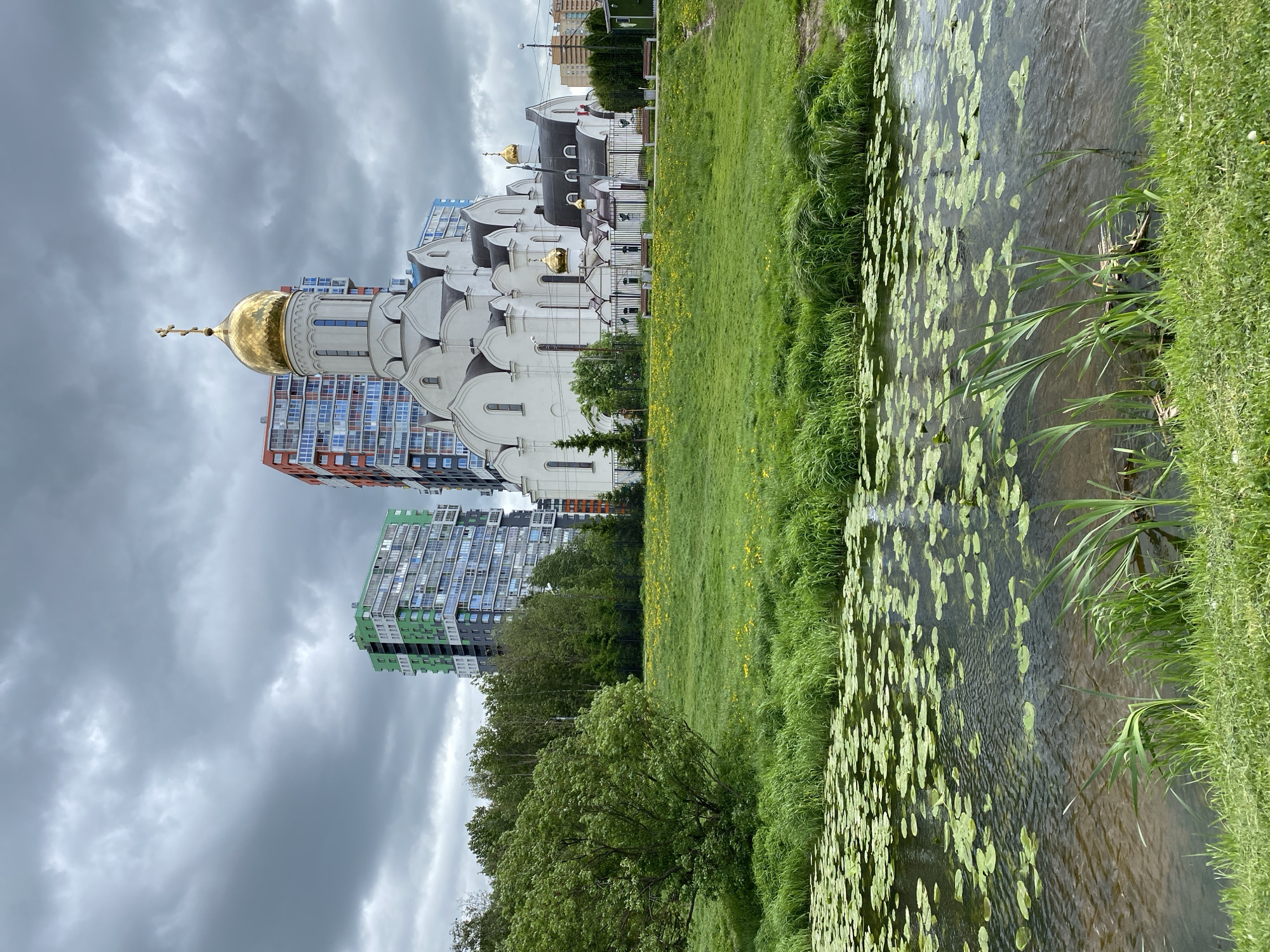 The image depicts a scene in an urban environment. In the foreground, there is a body of water with numerous lily pads floating on the surface. The water is bordered by green grass and aquatic vegetation. Beyond the water, there is a grassy area.
In the background, there is a Russian Orthodox church with white walls and multiple gold domes. Adjacent to the church are modern apartment buildings with balconies. The sky is overcast with dark clouds. The location appears to be in the city of Unknown, Unknown.