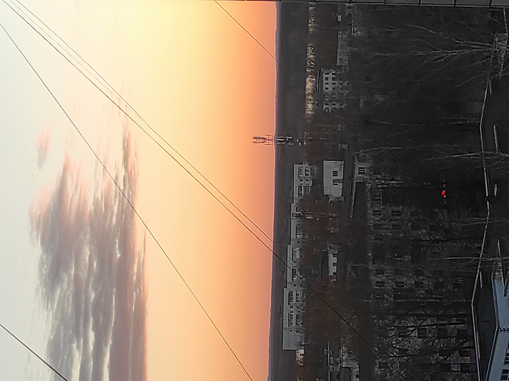 The image shows a view from a high vantage point in Chaikovsky, Russia, likely an apartment building. The scene is bisected vertically; the left side showcases a gradient sky, transitioning from a pale white near the horizon to a deep orange at the top, with wispy clouds. Power lines stretch diagonally across this part of the image. The right side of the image depicts the cityscape: densely packed buildings, bare trees, and hints of greenery. In the center of the image, near the horizon line, a radio tower stands. A faint red light appears near a building in the darker area.