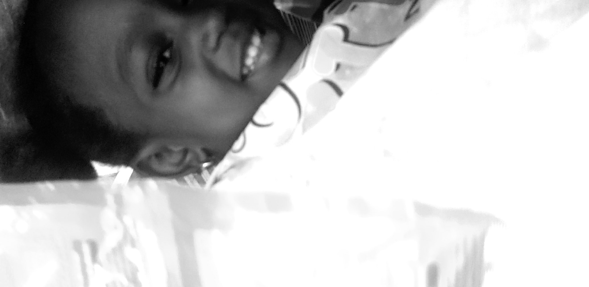 A black and white, close-up photograph shows a child's face peeking out from behind a large, blurry, white object. The child, who appears to be of African descent, is smiling and looking towards the camera. The child's hair is dark and short. The child is wearing a light-colored garment with a patterned design. The background is dark and out of focus. The image is cropped tightly, with the child's face occupying the right side of the frame. The white object in the foreground is so blurred that it is impossible to identify its nature or purpose. The overall impression is one of warmth and happiness, despite the lack of detail in the surrounding environment. The image is not specific to Narayi, Nigeria.