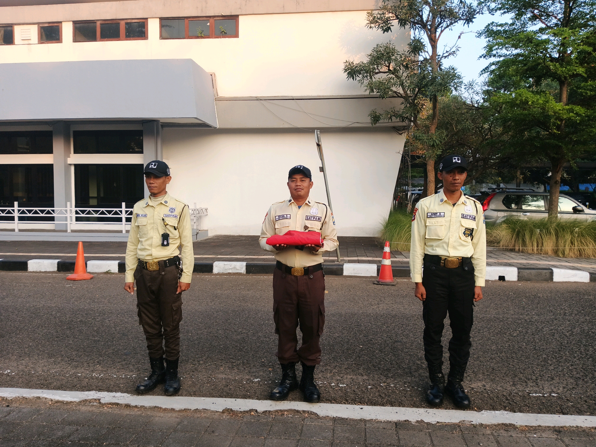 Three security guards stand in formation outdoors in front of a building. The guard on the left wears a tan uniform and a black baseball cap with a logo. The guard in the center wears a similar tan uniform and black cap, and holds a folded red cloth in his hands. The guard on the right also wears a tan uniform and black cap. All three guards wear dark pants and black boots. They stand on asphalt with a white and black striped curb to their left and a small orange traffic cone to the left of the leftmost guard. A white fence is visible behind the guards, and the building facade is a light color with rows of dark windows. Trees and other vegetation are to the right of the guards. The setting appears to be daytime, with the sun casting shadows. The scene is likely in Bandung City, Indonesia, given the context provided.