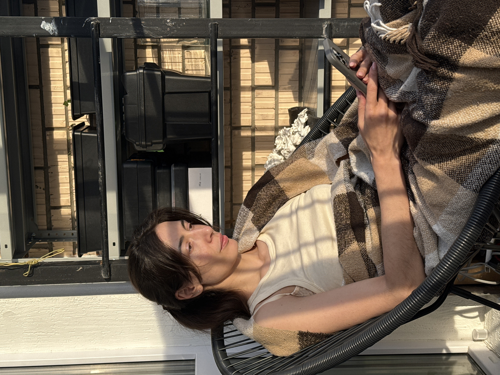 A person is seated outdoors on a black, wire-frame chair and covered with a brown-and-beige plaid blanket with tassels. The person holds a cell phone in their hands, which are positioned in front of them over the blanket. The individual wears a beige, ribbed tank top. The location appears to be a balcony, and there are black equipment cases stacked vertically against a railing. The railing is also black. Behind the railing are exterior walls. The image is taken in the daytime, with strong sunlight creating shadows. The location cannot be determined as Perm, Russia.