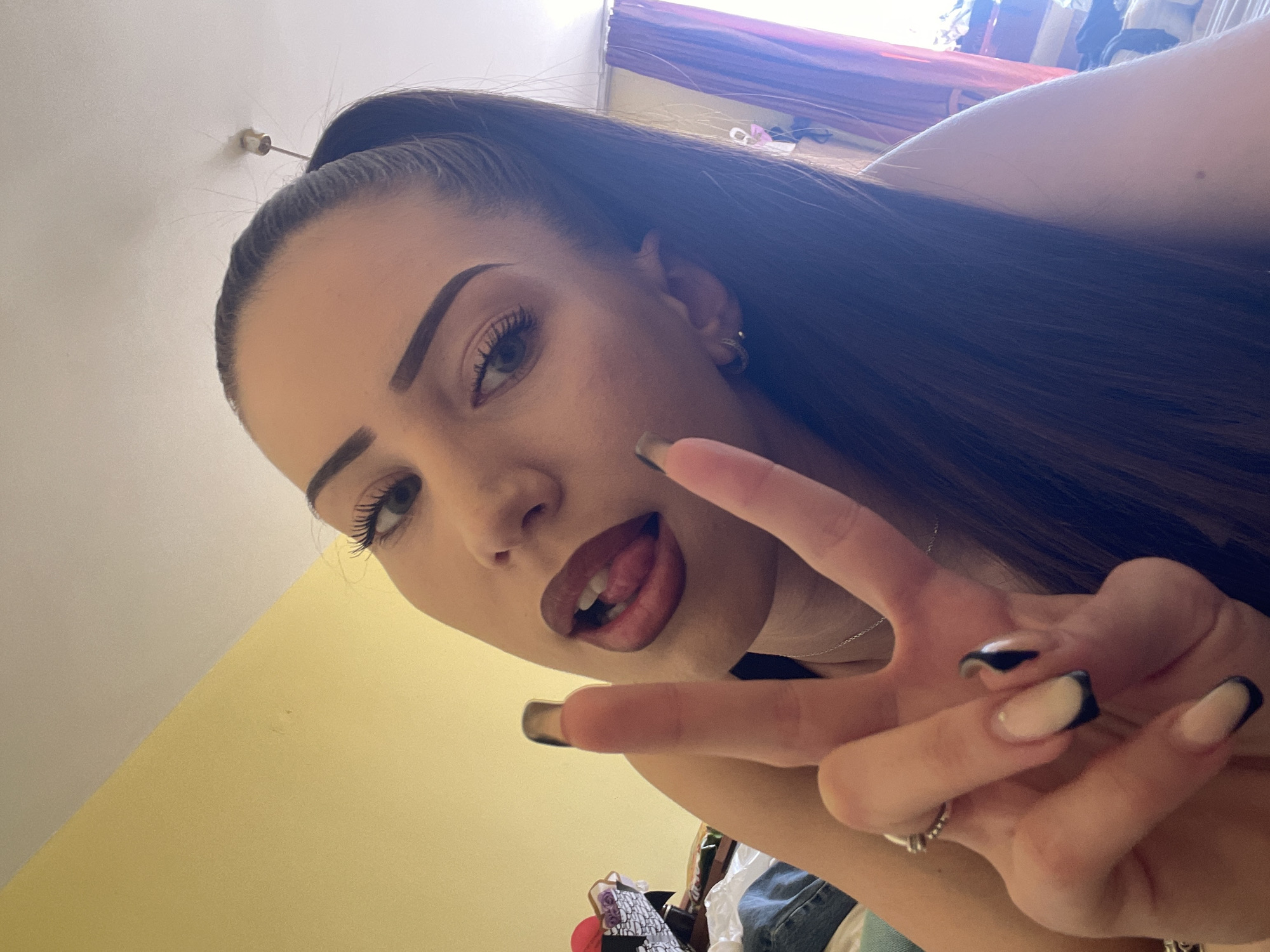 A close-up, upward-angled selfie captures a young woman with long, dark hair pulled back into a high ponytail or bun. She has prominent, styled eyebrows, dark eye makeup with noticeable eyelashes, and her lips are outlined in a darker shade with a lighter fill, slightly open with her tongue visible. She wears a small hoop earring in her left ear and a thin necklace. Her right hand is raised, making a "peace" or "V" sign, revealing long, square-shaped nails with a light base and black tips or outlines. A ring is visible on her right ring finger. The background is softly lit by natural light, showing a white ceiling with a small metal fixture and a light yellow wall. In the upper right corner, out-of-focus elements suggest a cluttered indoor setting, possibly with clothes and a reddish-purple curtain or fabric. The overall impression is casual and playful. There is no visible text.