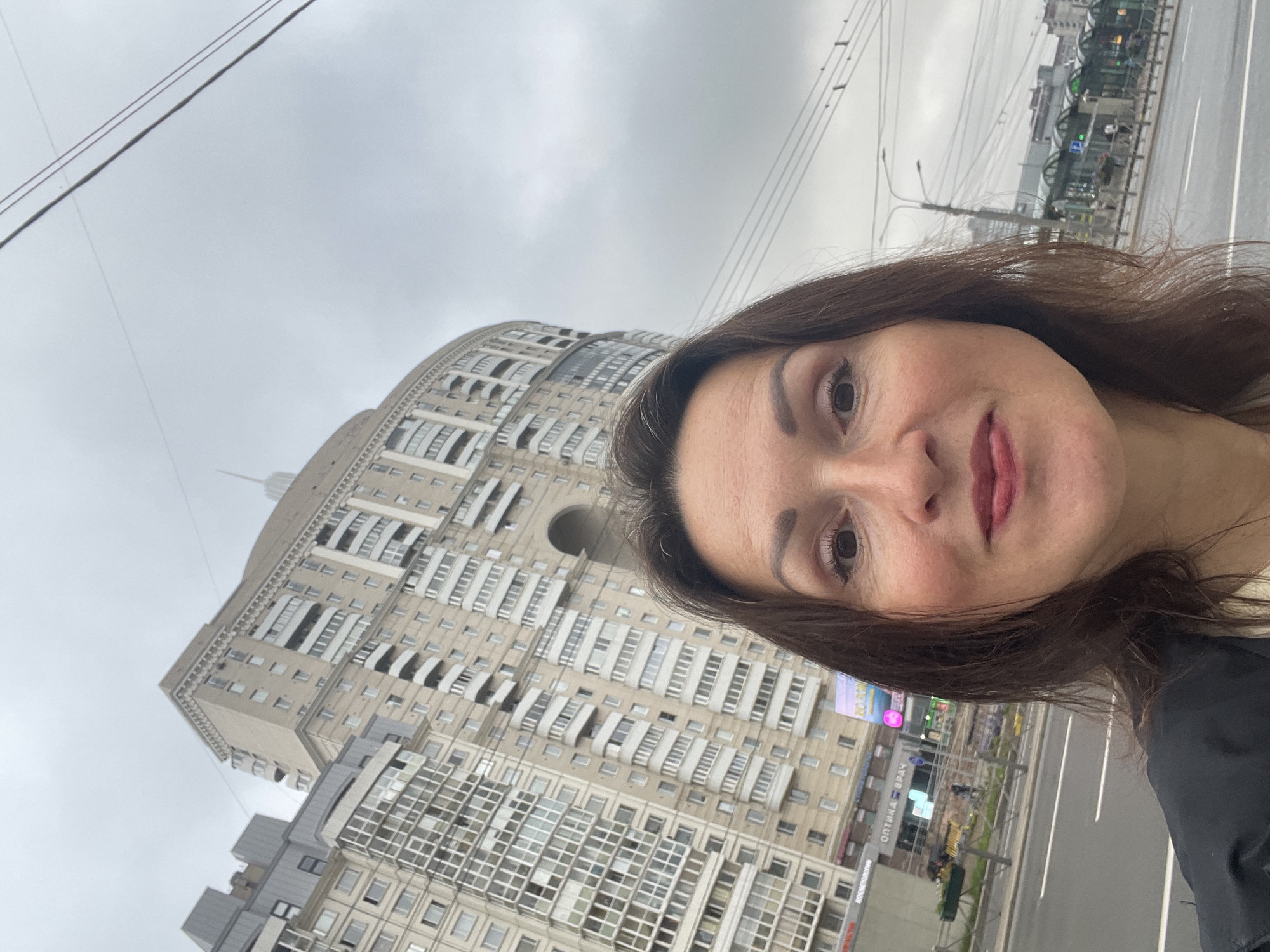A selfie is taken from a low angle in front of a large, curved apartment building with a pale yellow facade, many balconies, and arched architectural elements. The sky is overcast and gray. In the foreground, a woman with brown hair, wearing a black jacket, looks towards the camera with a slight smile. To the left of the building, a sign for "ОПТИКА ВРАЧ" (Optika Vrach - Optometrist Doctor) is visible, indicating a commercial space on the ground floor. The street in front of the building appears to be under some construction or renovation. Power lines are visible in the upper portion of the image, typical of urban environments. Further in the background, there are more buildings and what appears to be a public transport stop or station. The overall scene suggests an urban setting in Saint Petersburg, Russia, possibly in a residential area with commercial amenities.