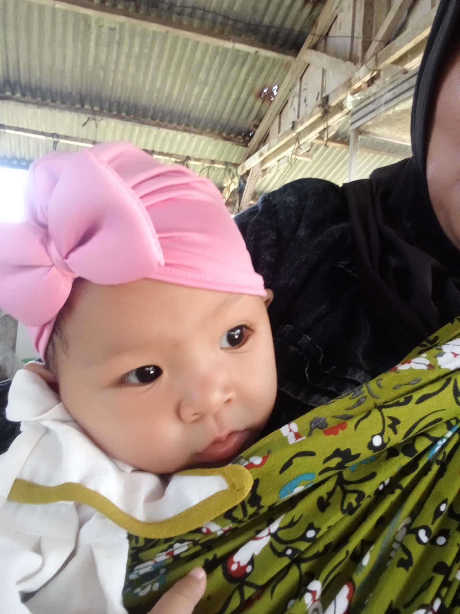 A close-up shot shows a baby wearing a pink turban with a bow, being held by a person wearing a black headscarf. The baby's face is partially visible, with large dark eyes and soft skin. The baby is dressed in a white top with yellow trim, and is being cradled in a green fabric with a floral pattern. In the background, a corrugated metal roof and wooden beams suggest an outdoor or semi-enclosed structure, possibly a dwelling or shelter in Negeri Lama, Indonesia.