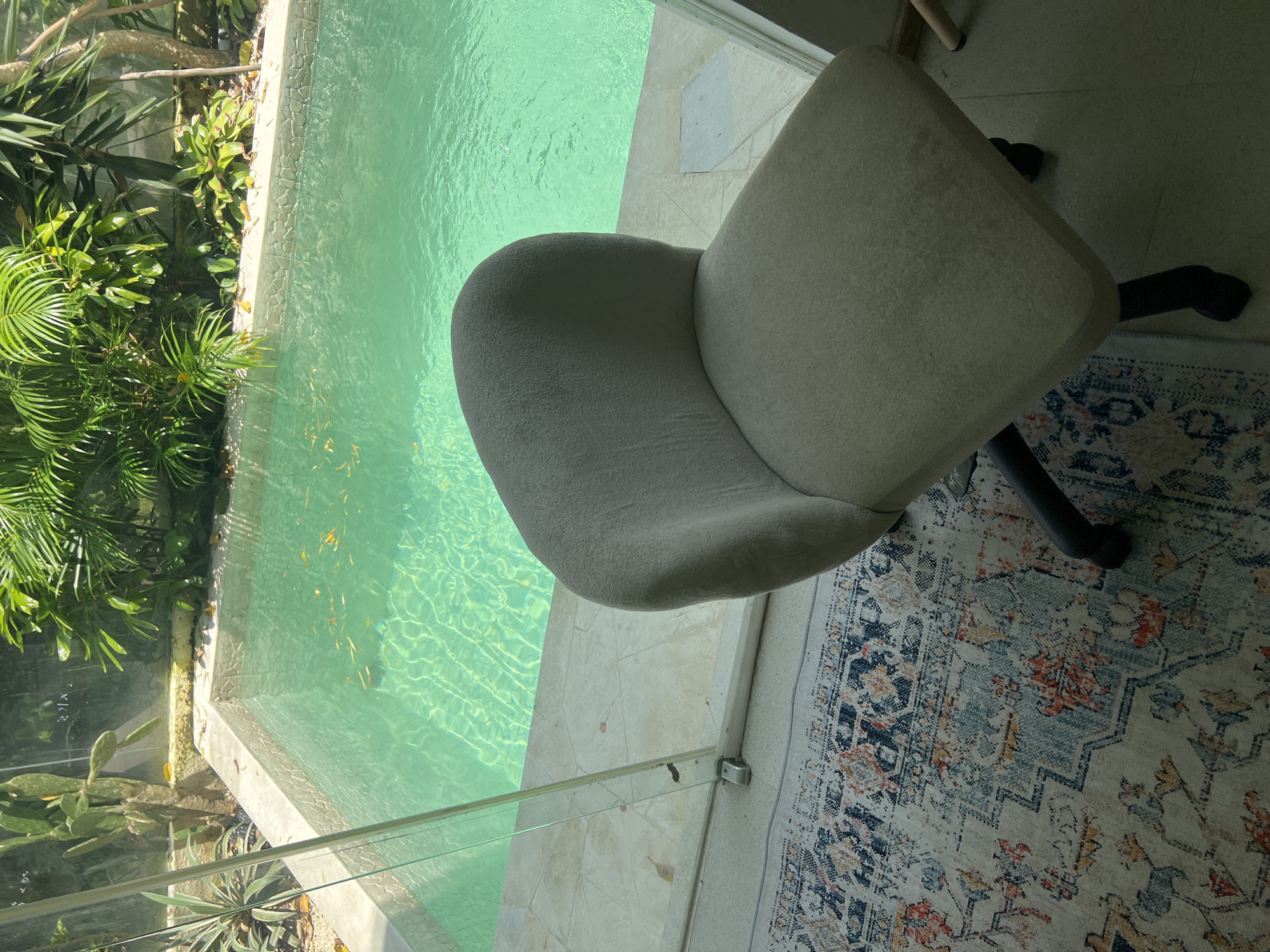 The image presents a high-angle view of an interior space in Buduk, Indonesia, bordering an outdoor pool area. The pool's turquoise water contains some floating leaves. Lush tropical vegetation is visible beyond the pool. A beige office chair with black legs and a five-star base sits on a patterned rug with blue, orange, and grey tones. A glass barrier separates the interior space from the pool. The floor is tiled with light-colored stone. The lighting suggests daylight conditions.