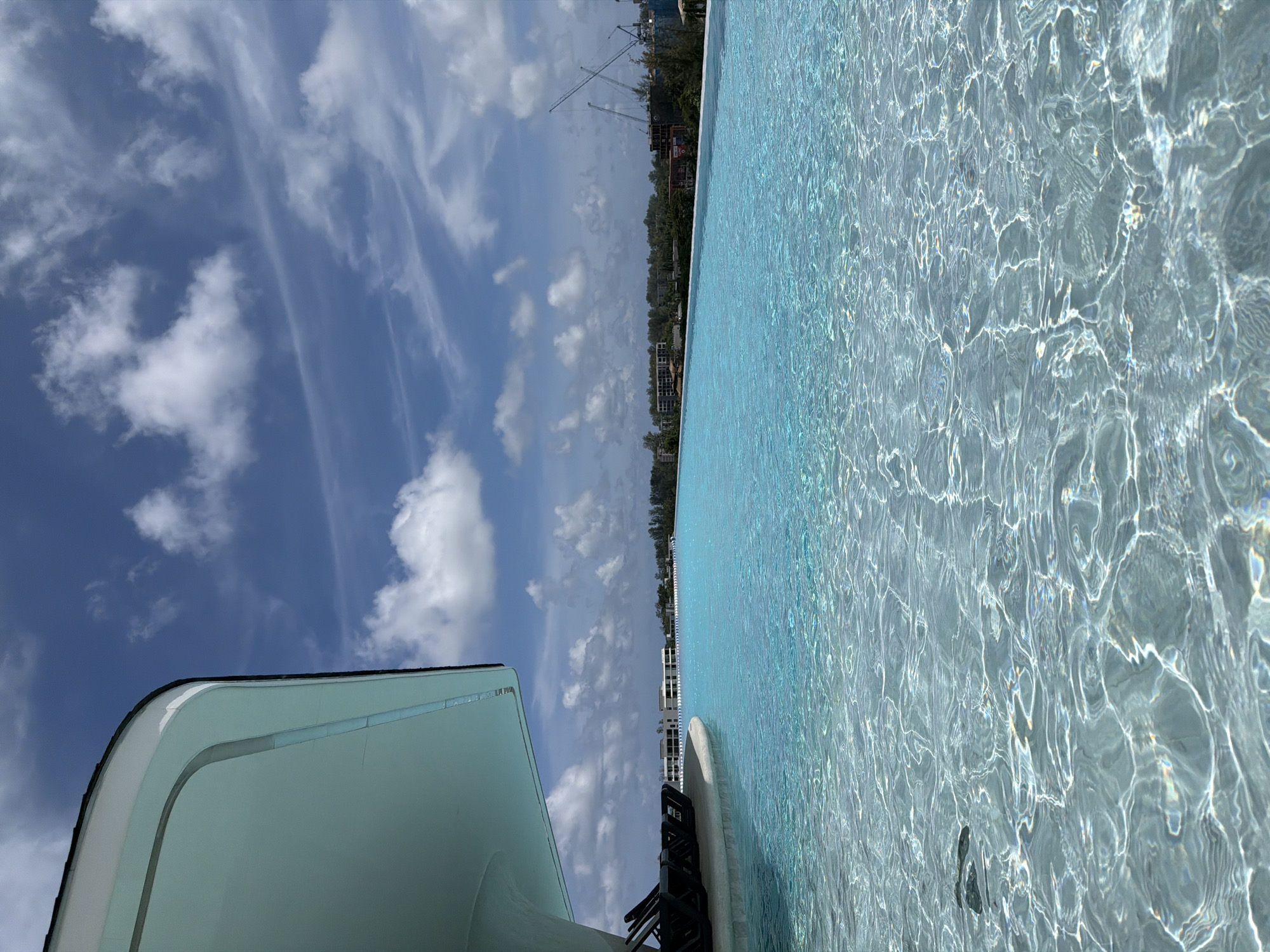 The image shows an infinity pool with clear turquoise water in Choeng Thale, Thailand. The pool's edge is visible in the foreground, with lounge chairs on the left side. The water is calm with sunlight reflecting off its surface, creating shimmering patterns. In the background, there is a line of buildings and lush green trees, with a crane visible in the distance, suggesting ongoing construction. The sky is blue with scattered white clouds.