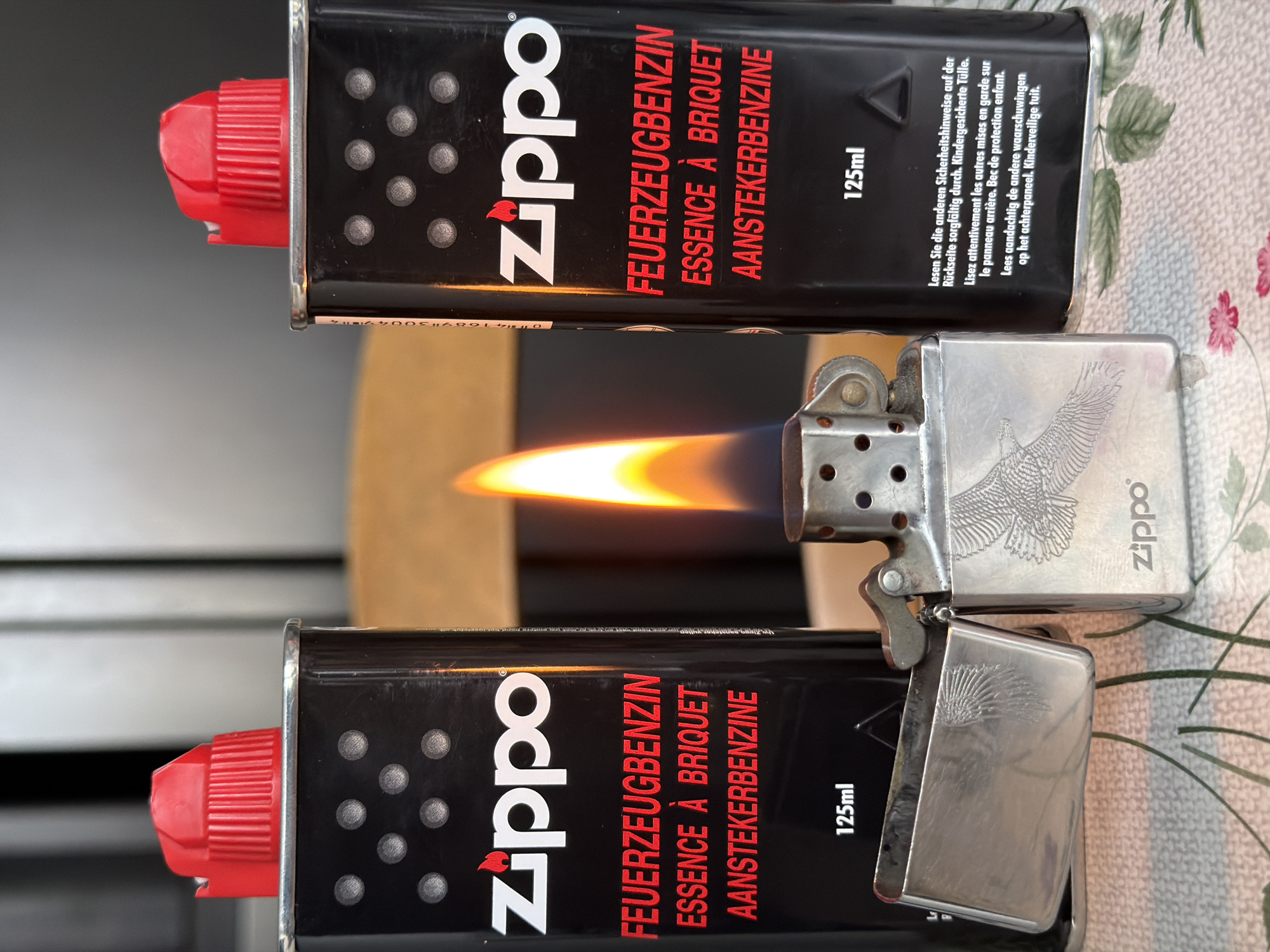 This image features two cans of Zippo lighter fluid, each with a red cap and the Zippo logo prominently displayed along with the product name in multiple languages: German (FEUERZEUGBENZIN), French (ESSENCE À BRIQUET), and Dutch (AANSTEKERBENZINE). The cans are labeled as containing 125ml of fluid. In the middle of the image, a Zippo lighter is actively burning with a bright yellow flame. The lighter has an eagle design engraved on its surface. The background is a patterned surface, possibly a tablecloth. There is no indication that the scene is located in Esch-sur-Alzette, Luxembourg.