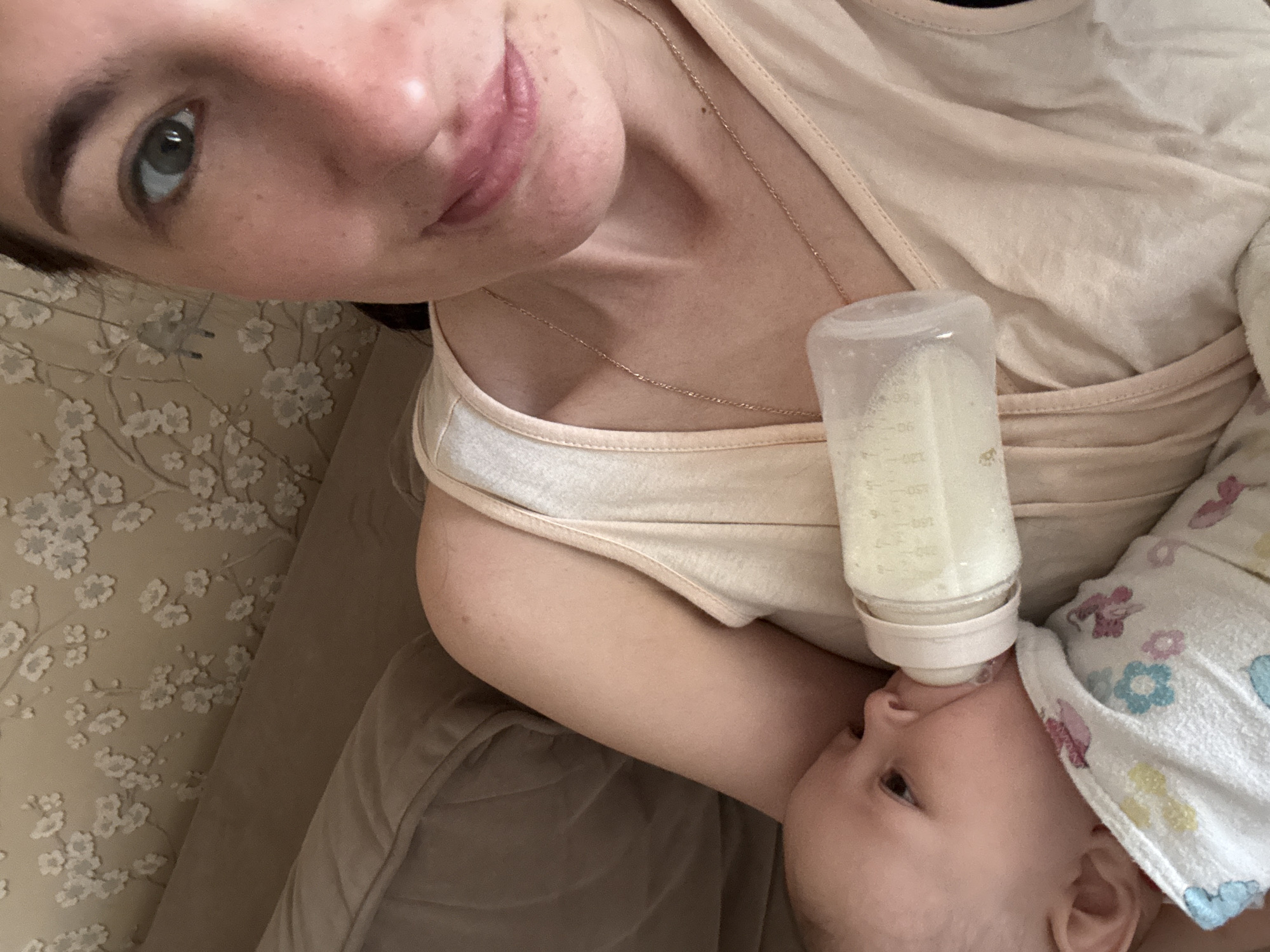 A woman lies in an indoor setting, holding a baby in her arms and feeding it from a bottle. The baby wears a light-colored cloth with floral patterns on its head. The environment appears to be a residential interior. The image does not show a location in the city of Sochi, Russia.
