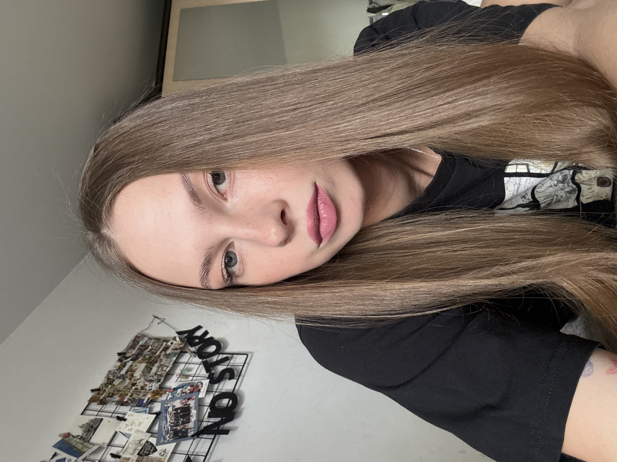 A young woman with long, straight blonde hair, fair skin, and light eyes is pictured in a medium shot, looking directly at the camera. She is wearing a black t-shirt. Behind her, on a light gray wall, is a black grid display featuring numerous photographs and postcards. To the left of the grid, black 3D letters spell out "NO STORY". The overall impression is an indoor setting, likely a bedroom or living space. There is no specific indication of the city of Perm, Russia, within the image itself.