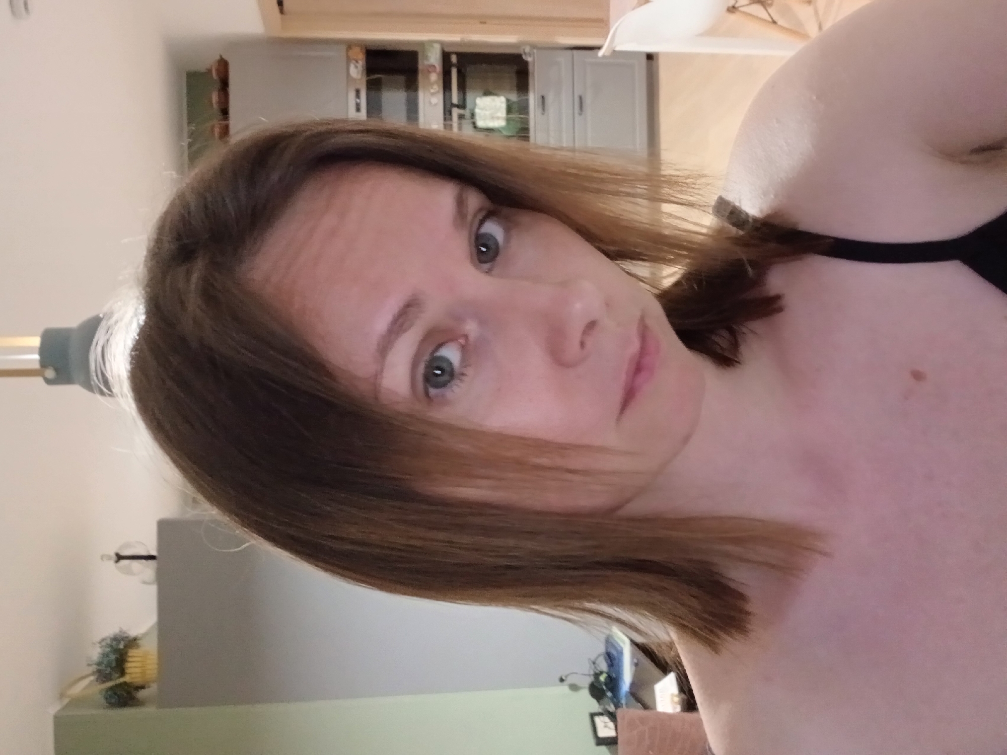 The image is a close-up, front-facing selfie of a woman with shoulder-length, reddish-brown hair, taken indoors. She is wearing a black strap, likely from a tank top or bra, visible on her right shoulder. Her skin appears fair with some faint freckles. The background suggests a domestic setting, possibly a kitchen or dining area, with grey cabinets and appliances visible. To the left of the woman, a desk with a computer monitor and a small framed picture can be seen. The lighting is moderately bright, creating soft shadows. The woman is looking directly at the camera with a neutral expression. The image does not provide any information about the city of Yekaterinburg, Russia, or any specific location within it.