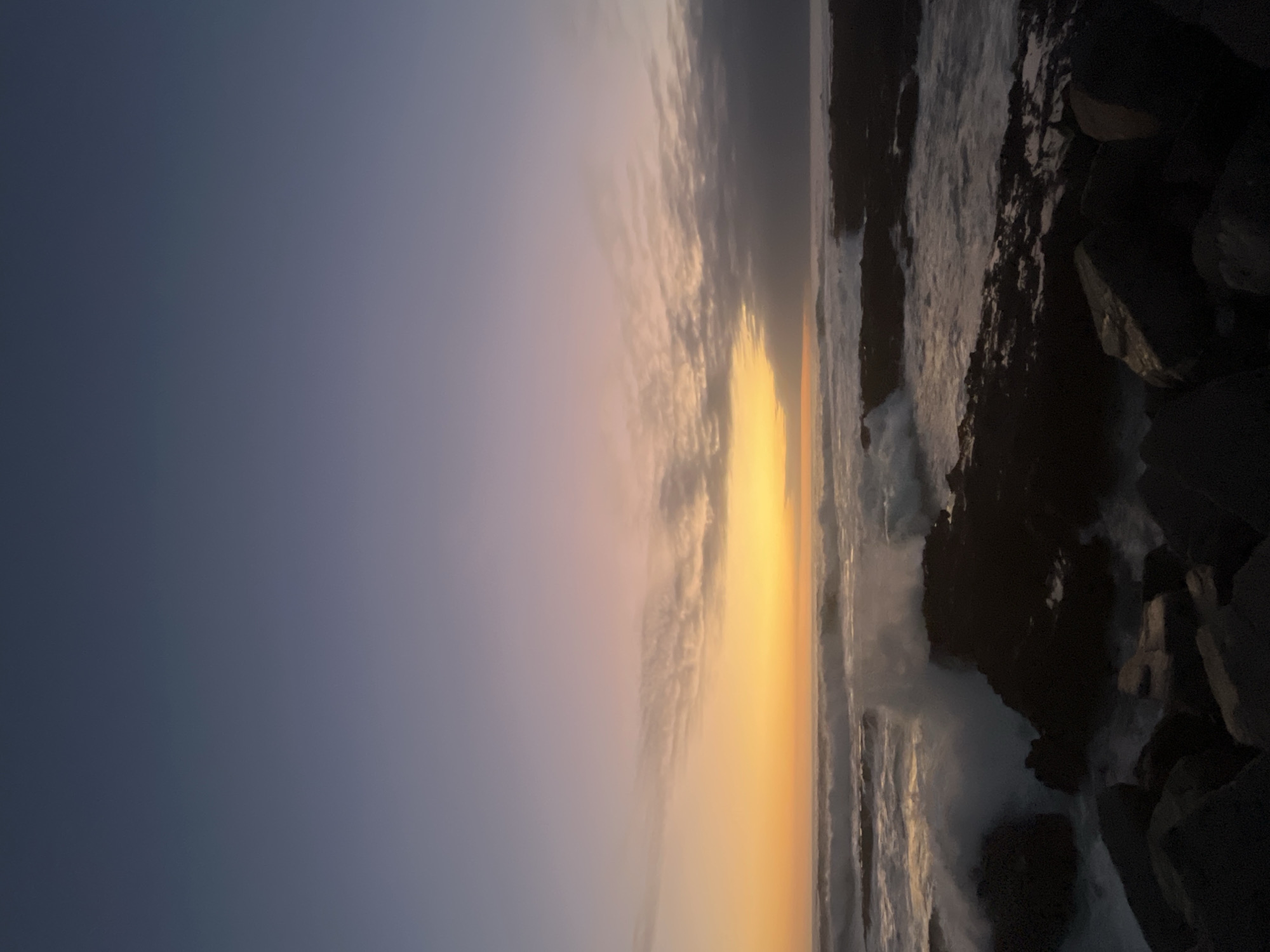 This is a serene landscape photograph depicting a coastal scene during either sunrise or sunset. The image, which appears to be rotated 90 degrees clockwise, shows a dramatic interplay of light and shadow across the sky, ocean, and rocky shoreline.
The sky, occupying the upper portion of the frame (when viewed horizontally), transitions in color. It begins with a deep, desaturated blue-grey at the top, gradually lightening and shifting to soft purplish-pink and pale blue tones closer to the horizon. Wispy, elongated clouds are scattered across the sky, subtly illuminated by the low light.
A prominent, intense golden-yellow glow emanates from the horizon line, indicating the precise location of the sun as it either rises or sets, casting a warm hue across the scene. This bright light is reflected in a shimmering path across the water.
The ocean fills the mid-ground, with active waves crashing and producing white foam, particularly evident as they meet the shore. The water itself appears dark and deep, contrasting with the radiant sky. On the right side of the frame, large, rugged rock formations or a jetty create a dark, silhouetted coastline. These rocks are mostly in shadow, though some areas catch glints of light from the crashing waves.
The scene conveys a sense of natural beauty and tranquility. The time of day is clearly dawn or dusk, marked by the distinctive gradient of colors in the sky and the low, golden light. There are no people, man-made objects, or visible text in the image.