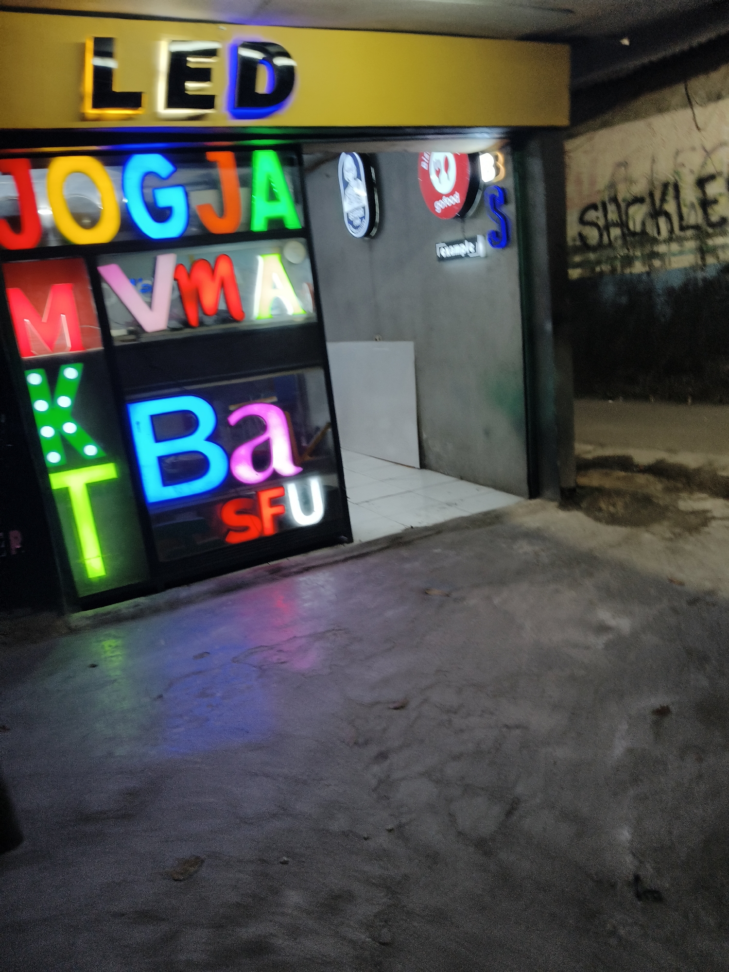 The image captures a storefront at night, featuring brightly lit LED signs. The main sign above the storefront reads "LED" in large, illuminated letters, with "L" in black and yellow, "E" in white and black, and "D" in blue and black. Below this, a row of colorful LED letters spells out "JOGJA" in red, yellow, blue, orange, and green. Further down, another set of LED letters reads "MM VA" in red, pink, and yellow. To the left, vertically aligned green and yellow LED letters spell out "MXT". To the right of "MM VA," a large blue LED letter "B" is prominent, followed by a pink "a," and below them, red and white LED letters spell "SFU." A circular red sign with white text is visible to the right of the "JOGJA" sign. The storefront itself appears to be a glass-enclosed area, possibly a shop or a kiosk. The ground in front of the store is paved and reflects the colorful lights from the signs. In the background, to the right, a wall is visible with graffiti that appears to say "SACKLE." The overall environment suggests an urban setting, likely a commercial street. The presence of the brand names and the style of the signage suggest this is a business selling or displaying LED signs. While the exact location within South Tangerang, Indonesia, cannot be determined from the image alone, the presence of "JOGJA" on the signage may refer to a location or a brand originating from Yogyakarta.