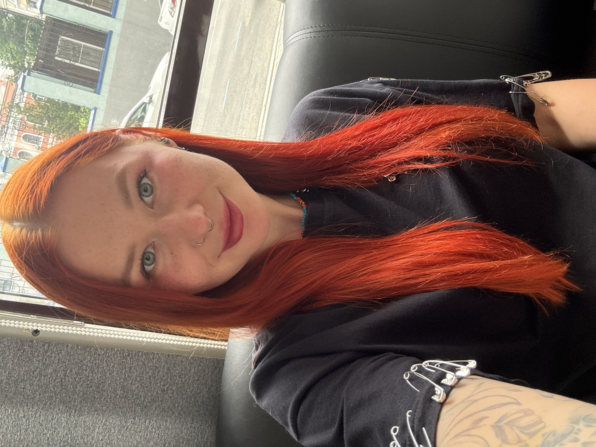 A woman with fair skin and bright red hair is seated indoors on a grey and black bus seat. Her green eyes are visible, and she has a nose ring. She is wearing a black long-sleeved shirt adorned with silver safety pins. A small beaded necklace is around her neck, and a portion of a tattooed arm is visible. Through the bus window, there is a glimpse of buildings with blue and white accents; however, the location cannot be determined.