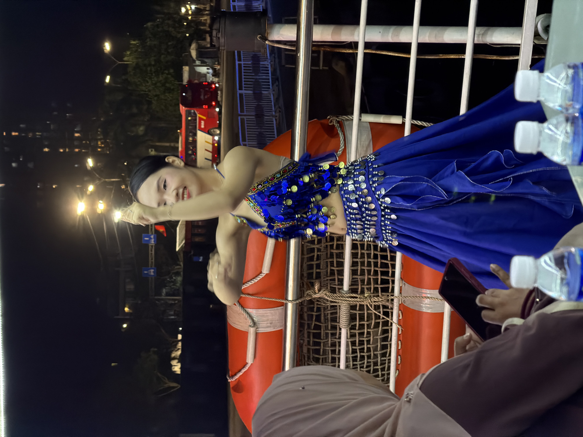 The image captures a vibrant nighttime scene, likely on a boat or pier in Đà Nẵng, Vietnam. The central subject is a young woman, possibly a performer, dressed in an elaborate two-piece blue costume. Her attire features a sequined and coin-embellished top and a flowing skirt, reminiscent of traditional or belly dance outfits. She has dark hair styled back and is smiling, striking a dynamic pose with one arm bent near her head and the other extended forward, suggesting she is performing, posing for a photo, or interacting with an audience.
In the foreground, partially visible on the bottom right, another person is seated, wearing a light-colored head covering and a dark top. They are holding a smartphone, seemingly recording or taking photos of the performer, and also have plastic water bottles nearby.
The setting is outdoors at night, illuminated by various artificial lights. Behind the performer, there are sturdy metal railings and an orange life preserver with rope, along with a mesh netting structure, indicating a marine or waterfront environment. In the distant background, streetlights glow, and a red and white bus, possibly with the text "PHÚ" visible on its side, can be seen along with other vehicles and buildings, hinting at an urban landscape alongside the water. The overall atmosphere suggests an evening of entertainment or a festive event by the waterfront.