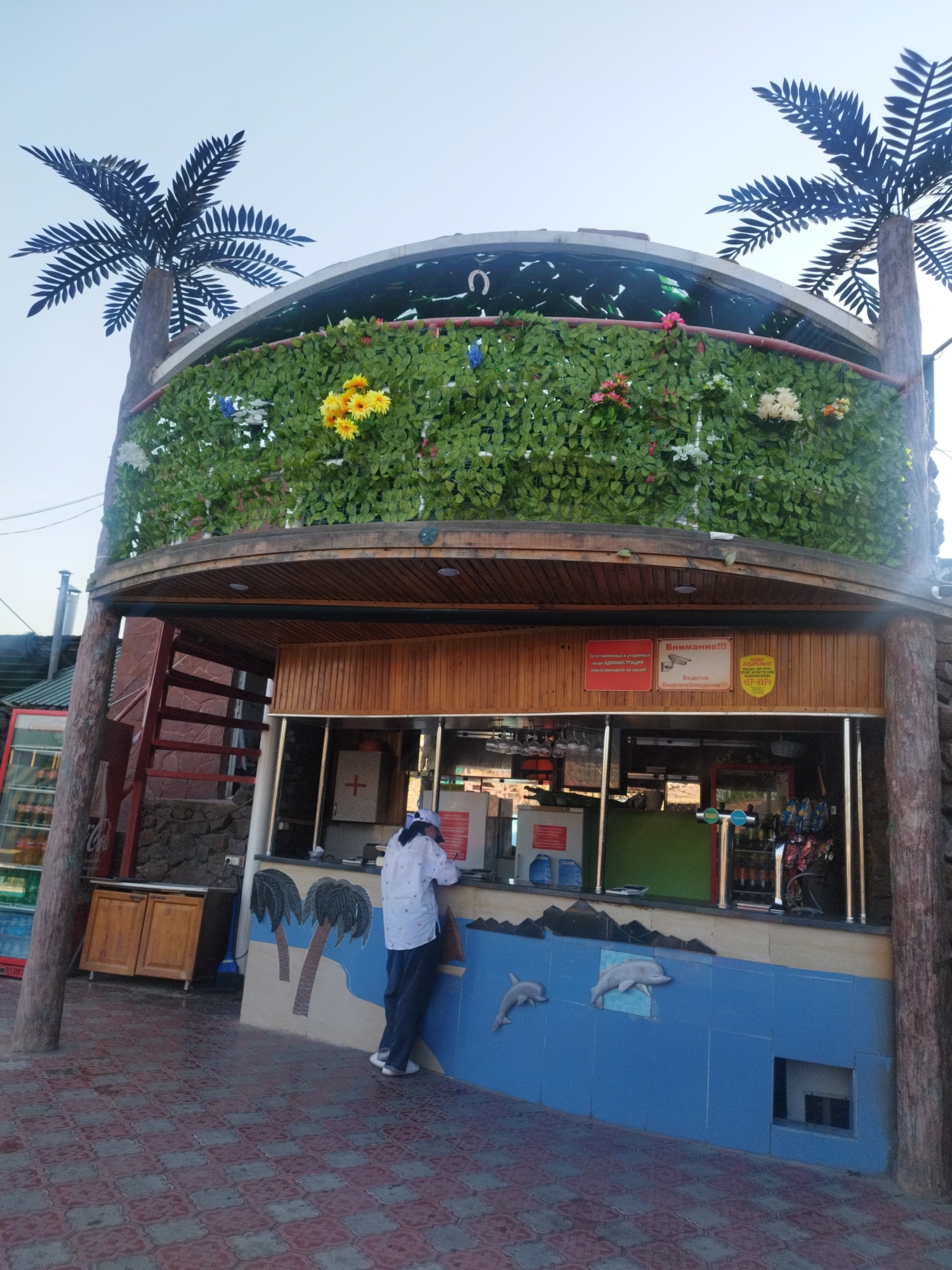 A man stands at a counter in front of a building adorned with palm trees and artificial greenery. The building appears to be an open-air establishment, possibly a bar or food service. Behind the man, there are signs in Russian, one reading "Внимание!!!" (Attention!!!) and another indicating video surveillance. The ground is paved with decorative tiles. The scene appears to be outdoors, with a clear sky overhead.