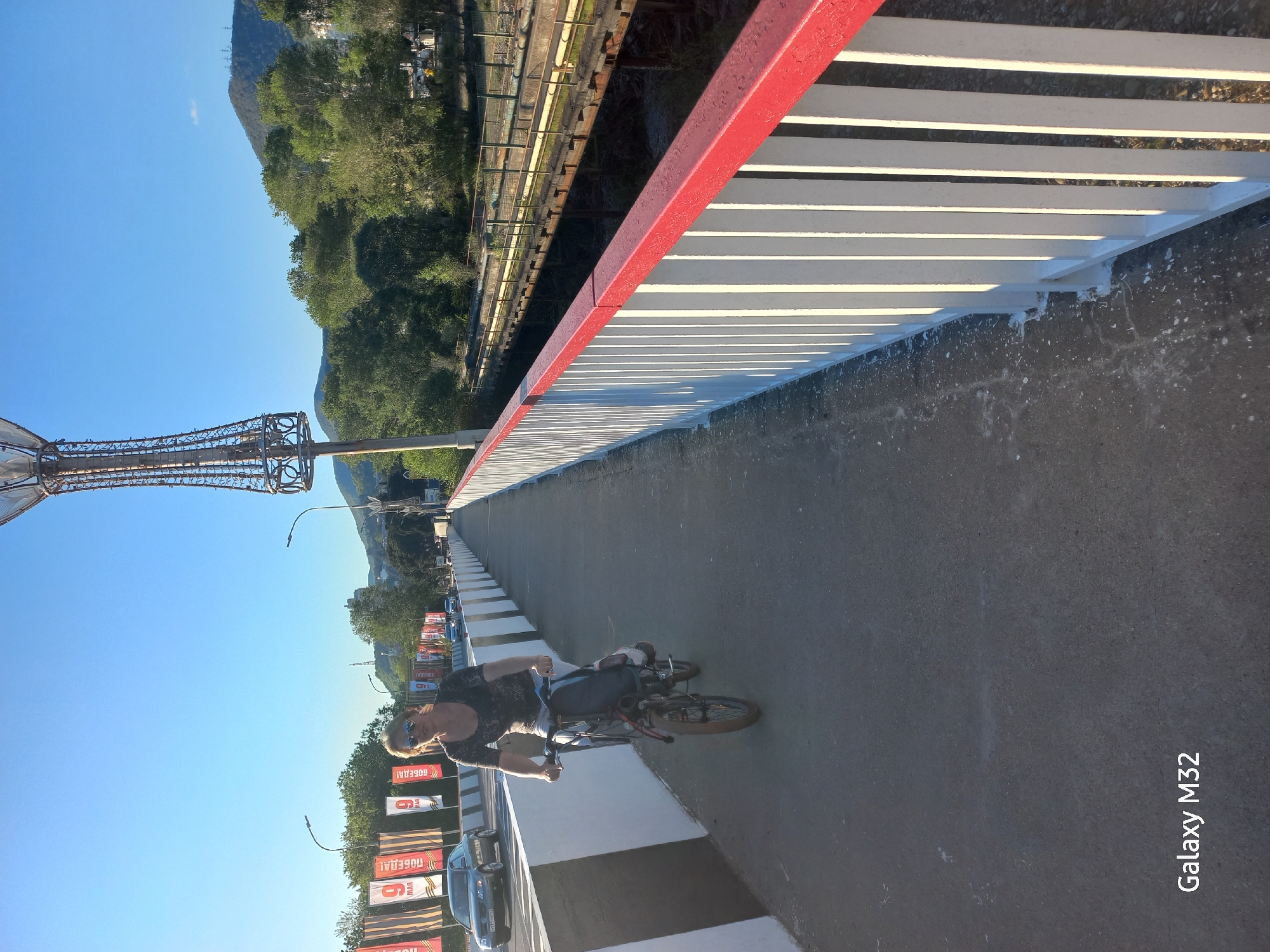 A woman is riding a bicycle on a concrete bridge sidewalk in Sochi, Russia. The bridge has white metal railings with a red top rail. Adjacent to the bridge, on the right side of the image, are railway tracks with trees. The cityscape is visible in the distance, with mountains in the background under a clear blue sky. On the left side of the image are cars, trees and signs with the text "ПОБЕДА!" and the number "9". The camera used to take the photo is a Galaxy M32.